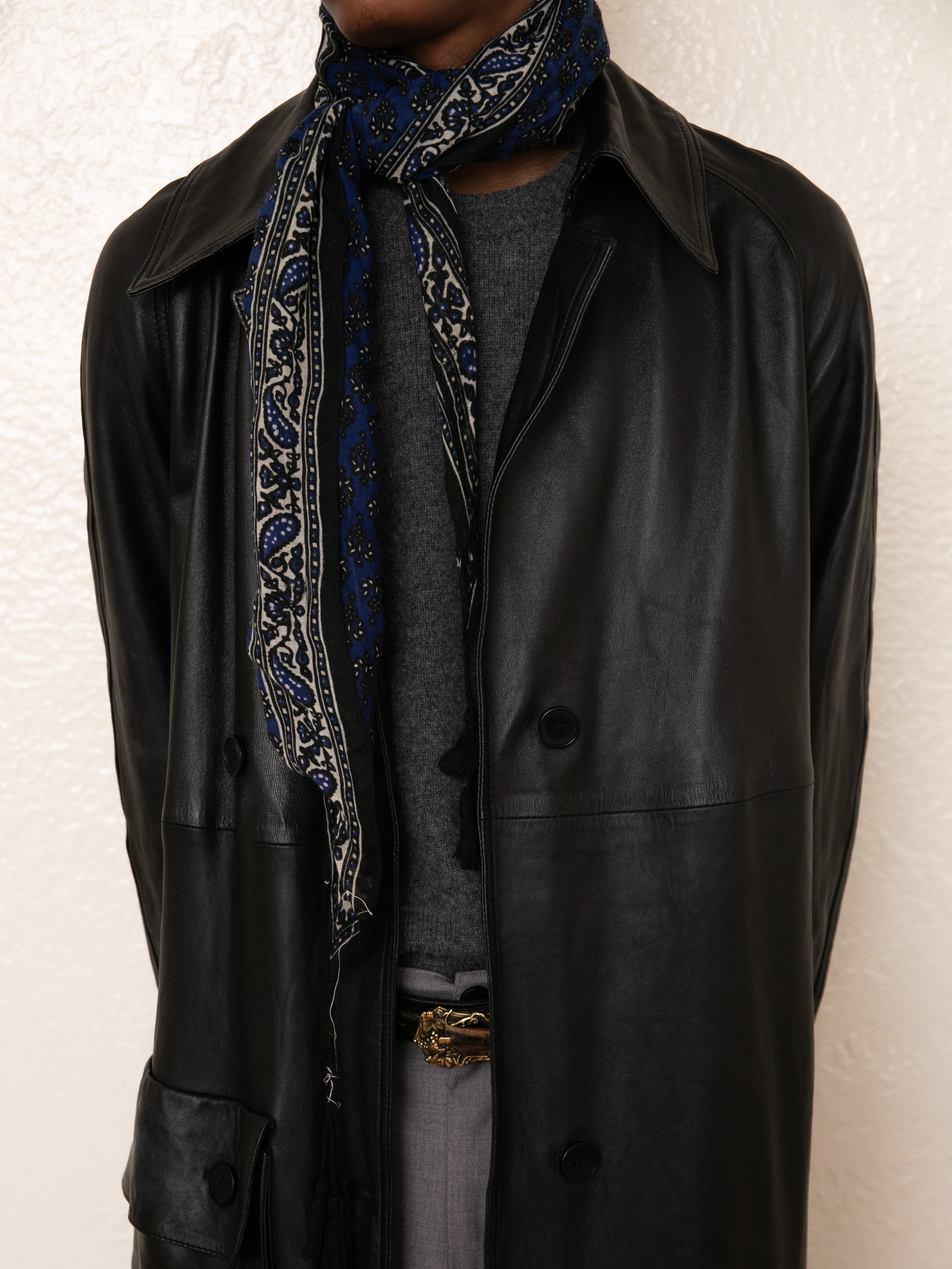 A person models FOUND's ethically crafted Leather Trench Coat with a gray sweater and blue patterned scarf against a textured white wall.