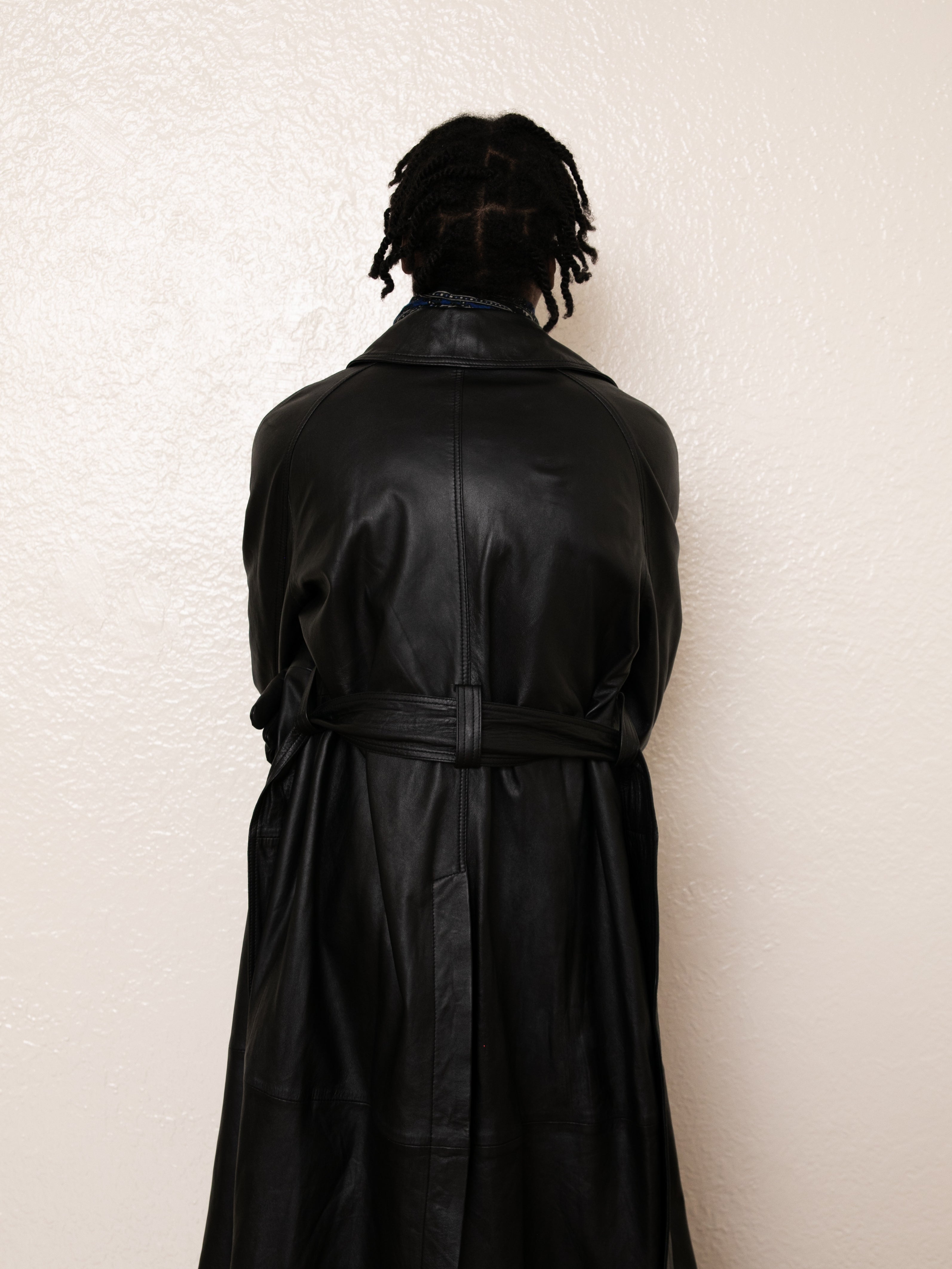 A person with braided hair, wearing the FOUND Leather Trench Coat in black, stands with their back to the camera against a plain white wall.