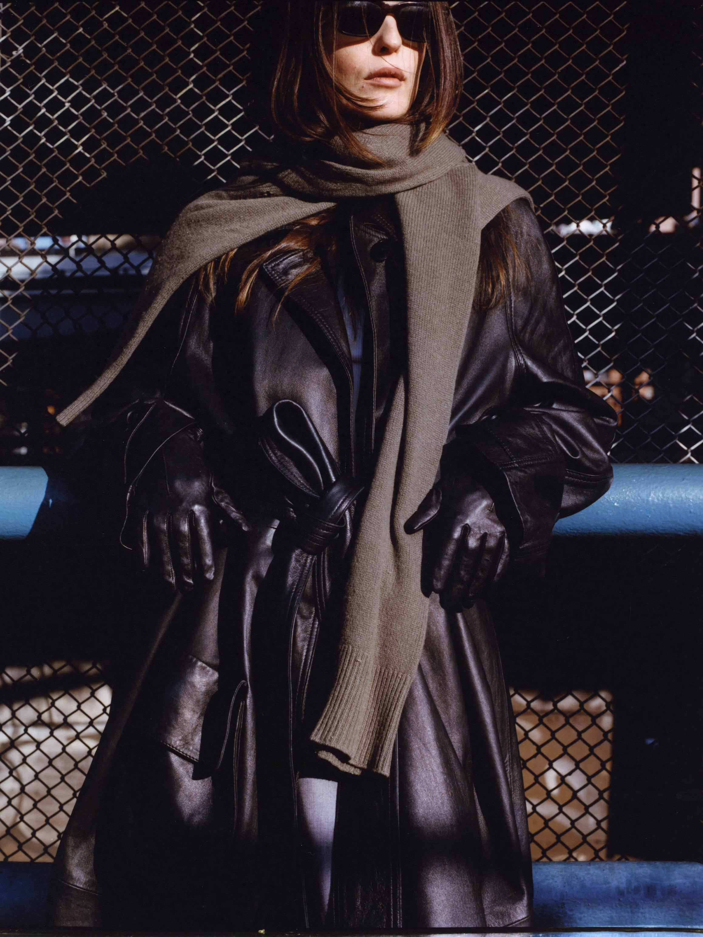 A person in a sleek leather trench coat by FOUND, showcasing exquisite cowhide craftsmanship, stands before a chain-link fence, complemented by a gray scarf and sunglasses, with gloves adding to the polished ensemble.