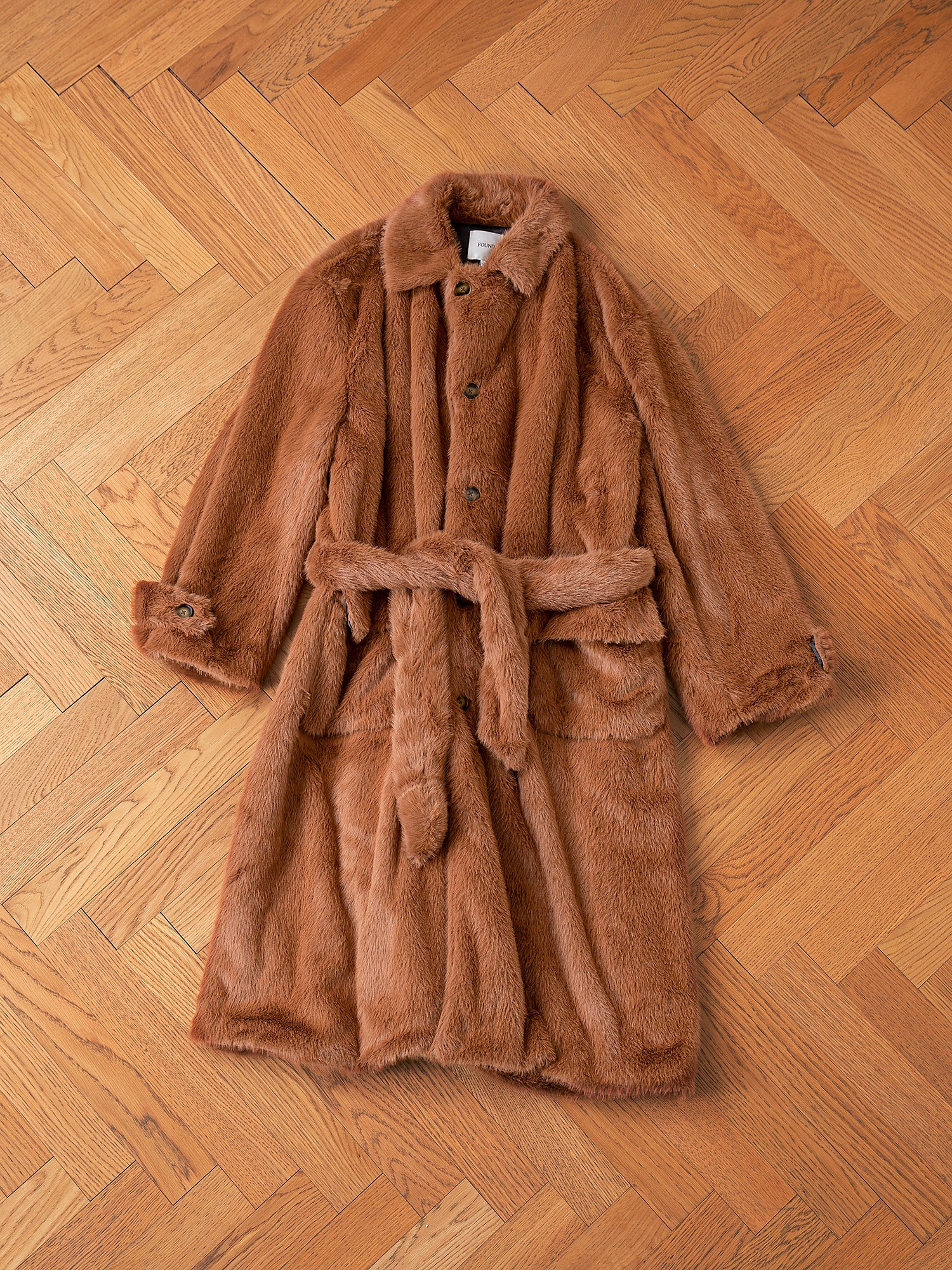 The Long Faux Fur Coat by FOUND, a stylish brown double-breasted piece with buttons and a waist belt, is elegantly laid out on a wooden parquet floor.