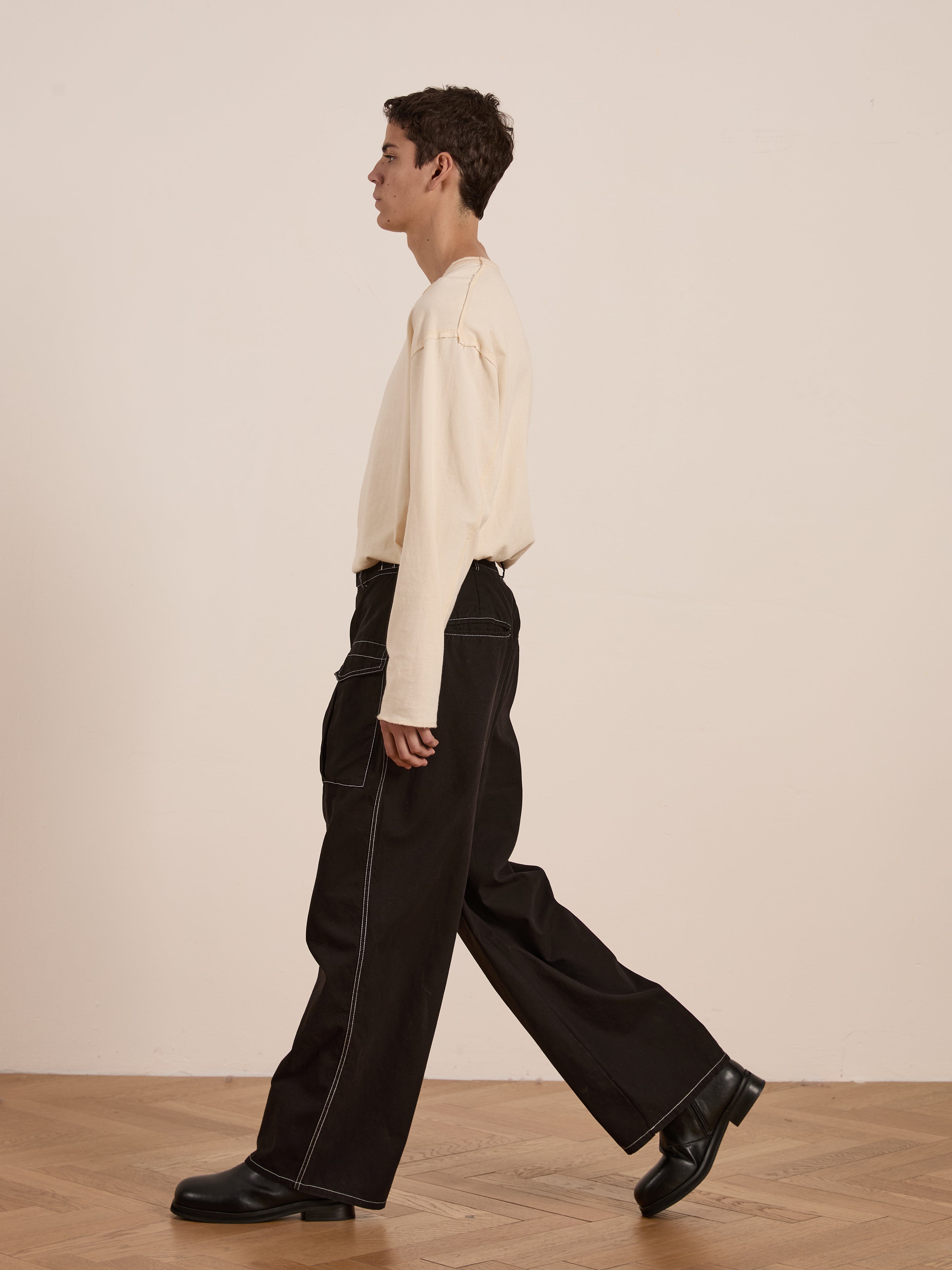 A person in a beige long-sleeve shirt and FOUND's Lucca Black Contrast Pocket Pants, featuring a relaxed straight-leg wide fit with white stitching, walks on a wooden floor, shown from the side.