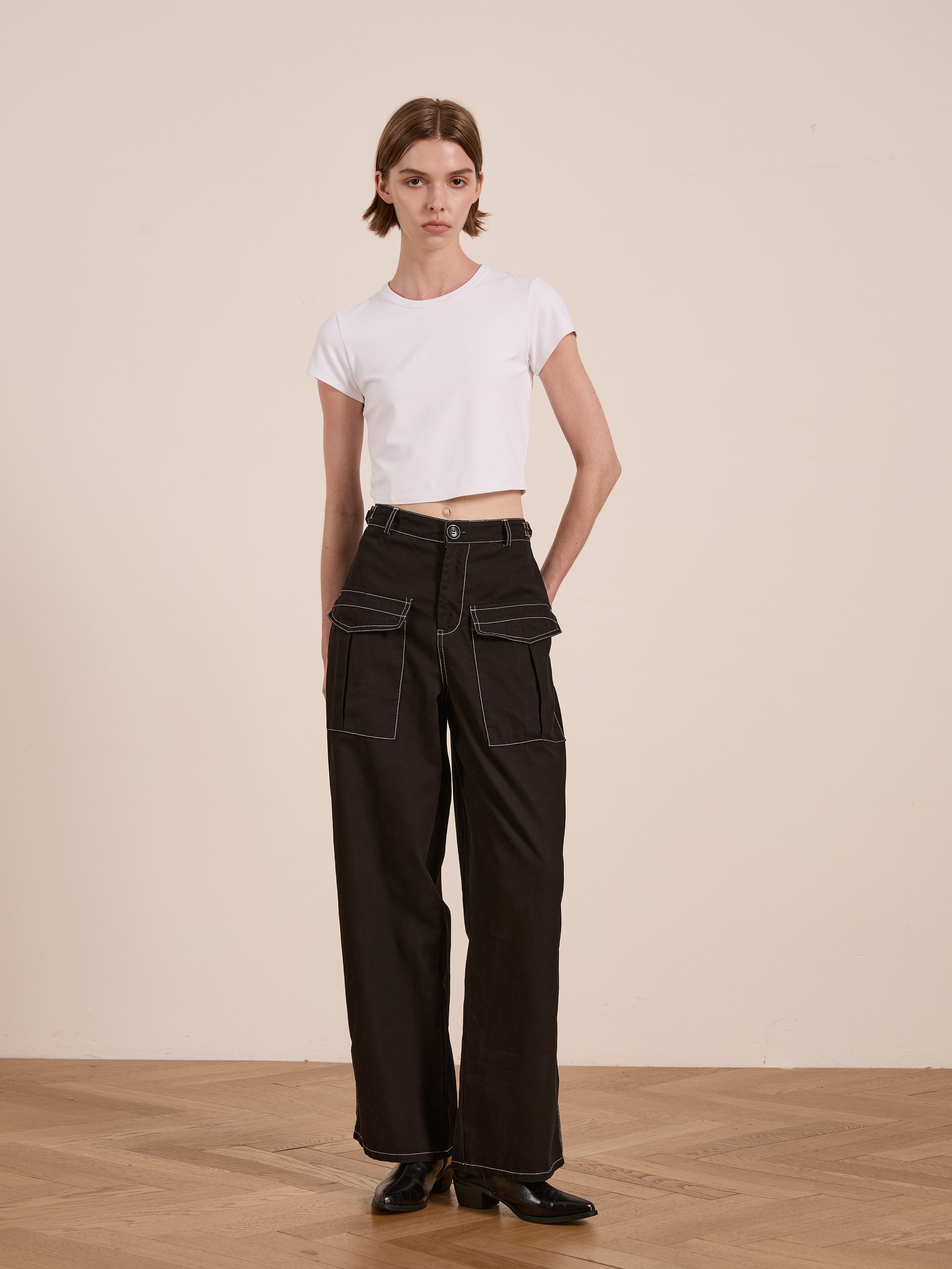 A person wearing a white crop top and FOUND's Lucca Black Contrast Pocket Pants with a relaxed straight-leg fit stands on a wooden floor against a plain wall.