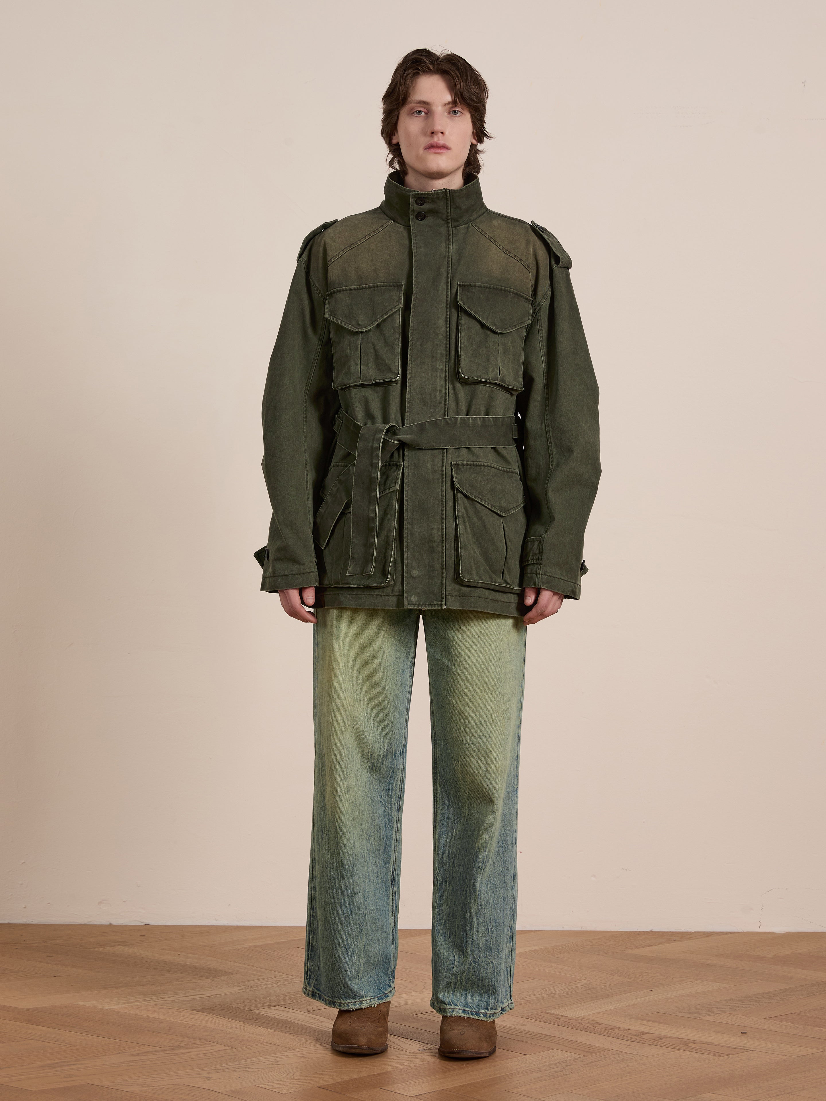 A person in a green FOUND M65 Utility Twill Pocket Jacket, with a relaxed fit, pairs it with wide-leg jeans and brown boots. Standing on wood flooring against a neutral background, this military-inspired outerwear exudes timeless style.