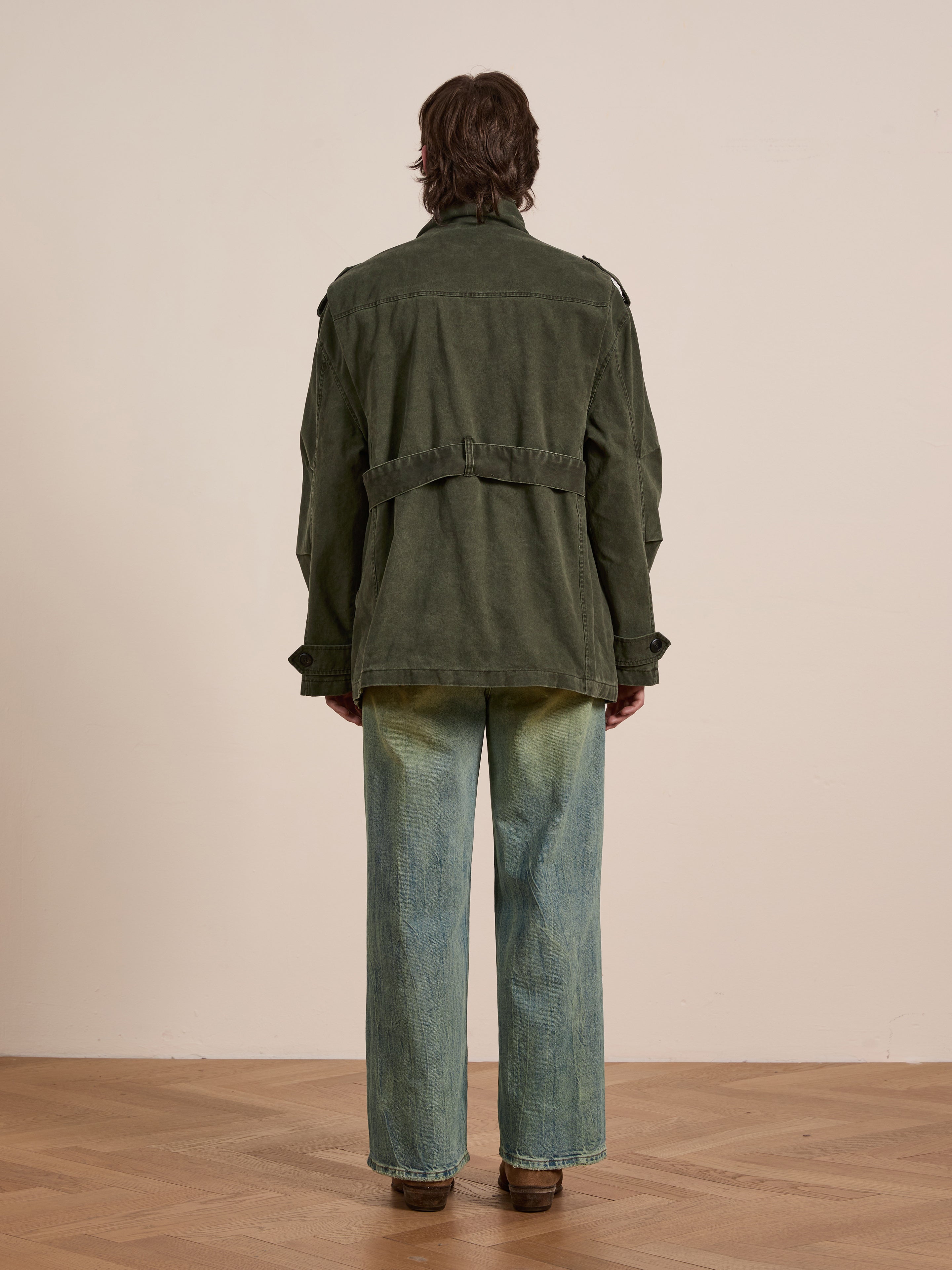 Someone wearing a FOUND M65 Utility Twill Pocket Jacket and faded blue jeans stands on a wooden floor, facing a blank wall.