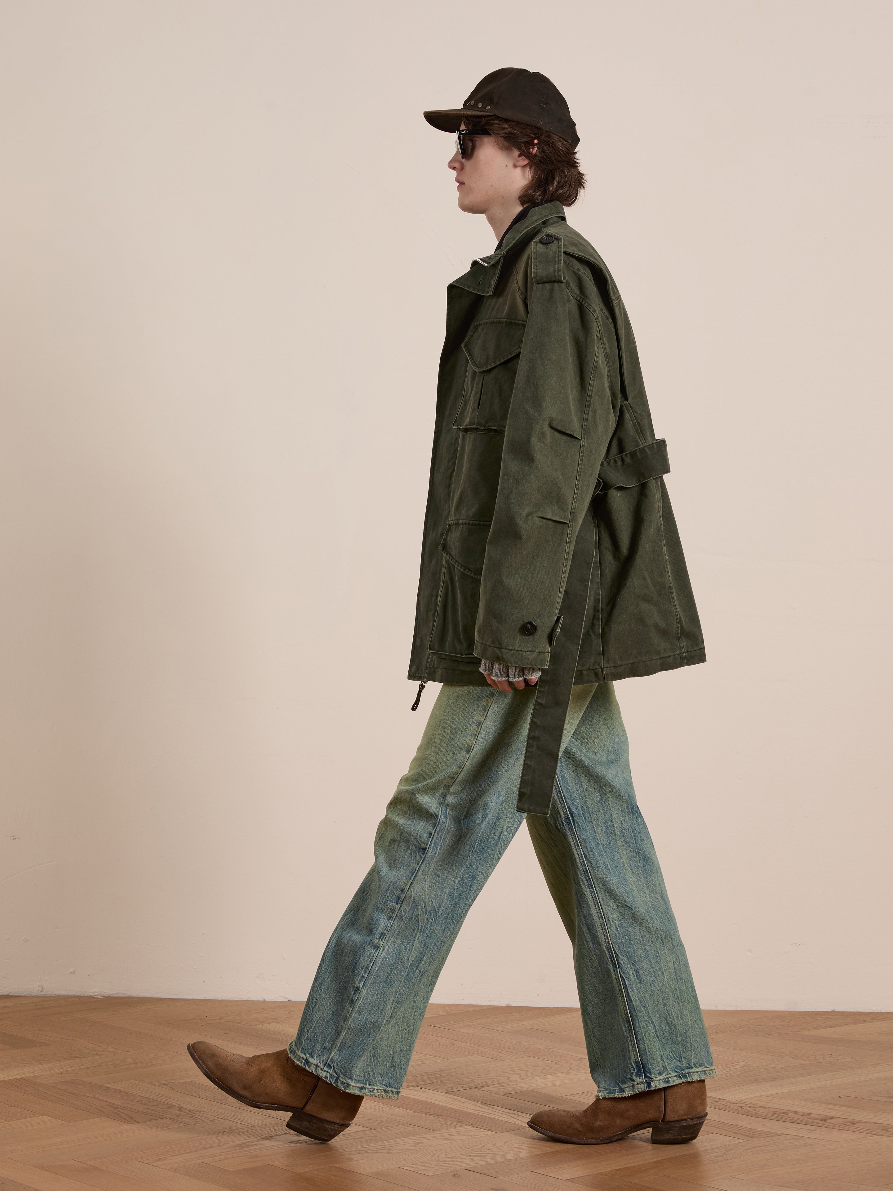 Wearing the FOUND M65 Utility Twill Pocket Jacket, jeans, and brown boots, a person strides sideways across a wooden floor with a dark cap and sunglasses.