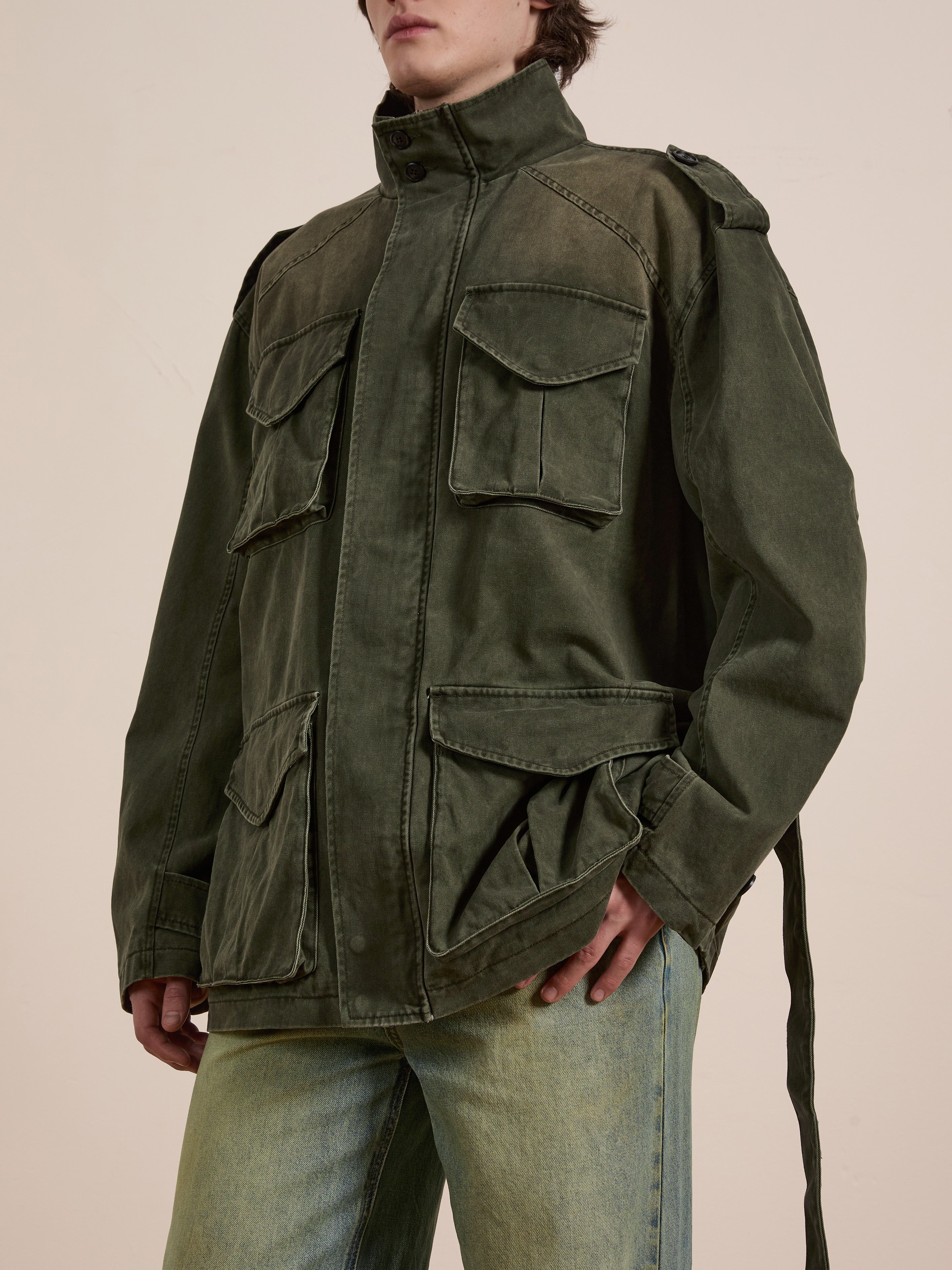 A person in a green M65 Utility Twill Pocket Jacket by FOUND, noted for its military-inspired style and large pockets, complements it with faded jeans. This unisex, relaxed-fit garment seamlessly integrates into their casual look.