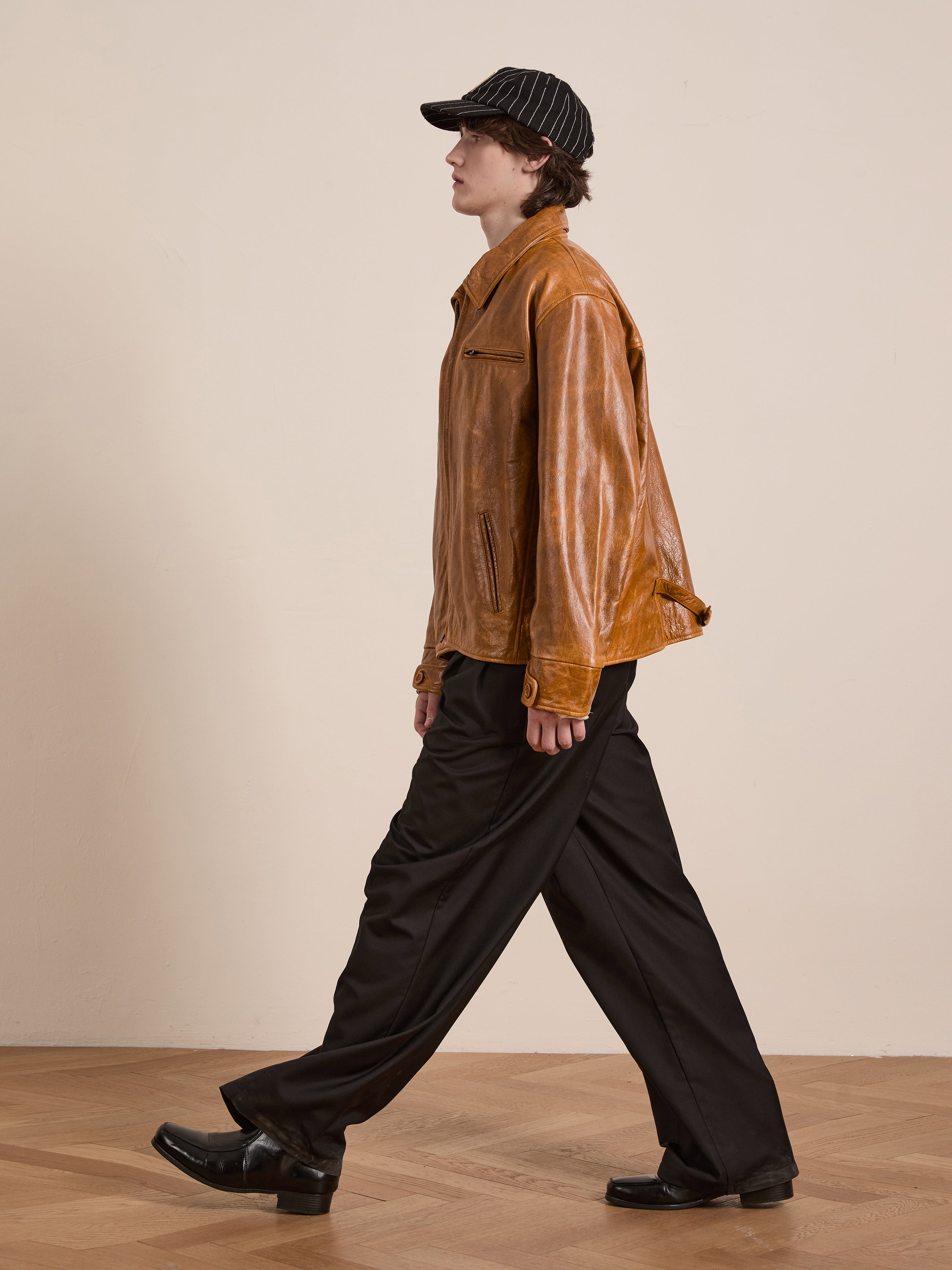 A person in a Marlboro Sun Faded Leather Racer Jacket by FOUND, black pants, and shoes strolls across a wooden floor, their look completed with a sleek black cap.