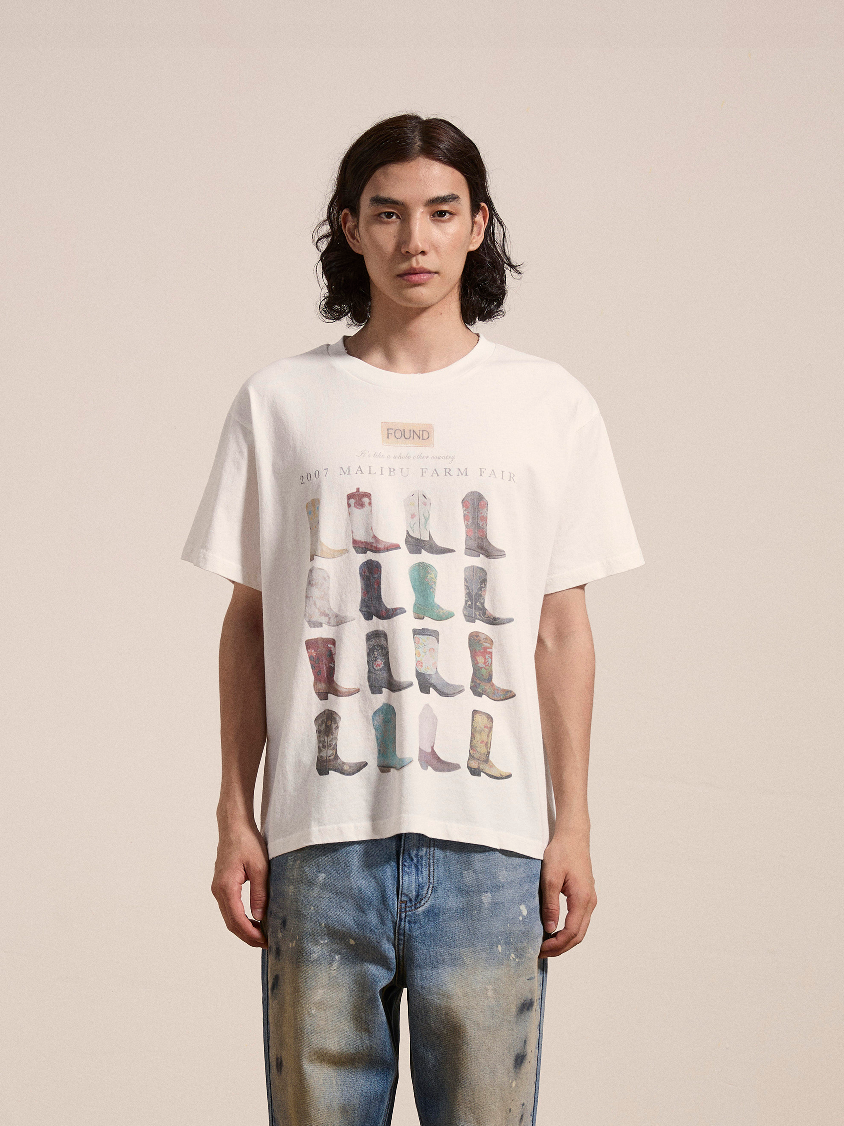 A person with long hair is wearing a Malibu Farm Fair Tee by FOUND, made from soft cotton and adorned with multiple boot images, while standing in distressed jeans against a plain background.