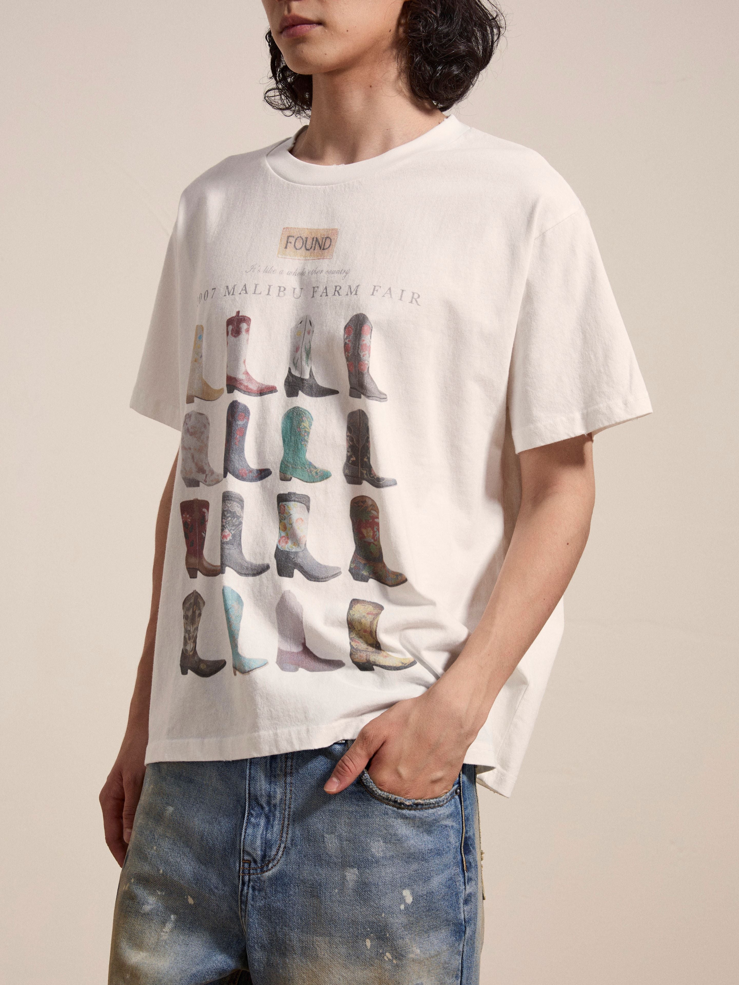 A person wearing a FOUND Malibu Farm Fair Tee made of 100% cotton, featuring an illustrated print of assorted cowboy boots and the text "1967 MALIBU FARM FAIR," has their hand in the pocket of their blue jeans.