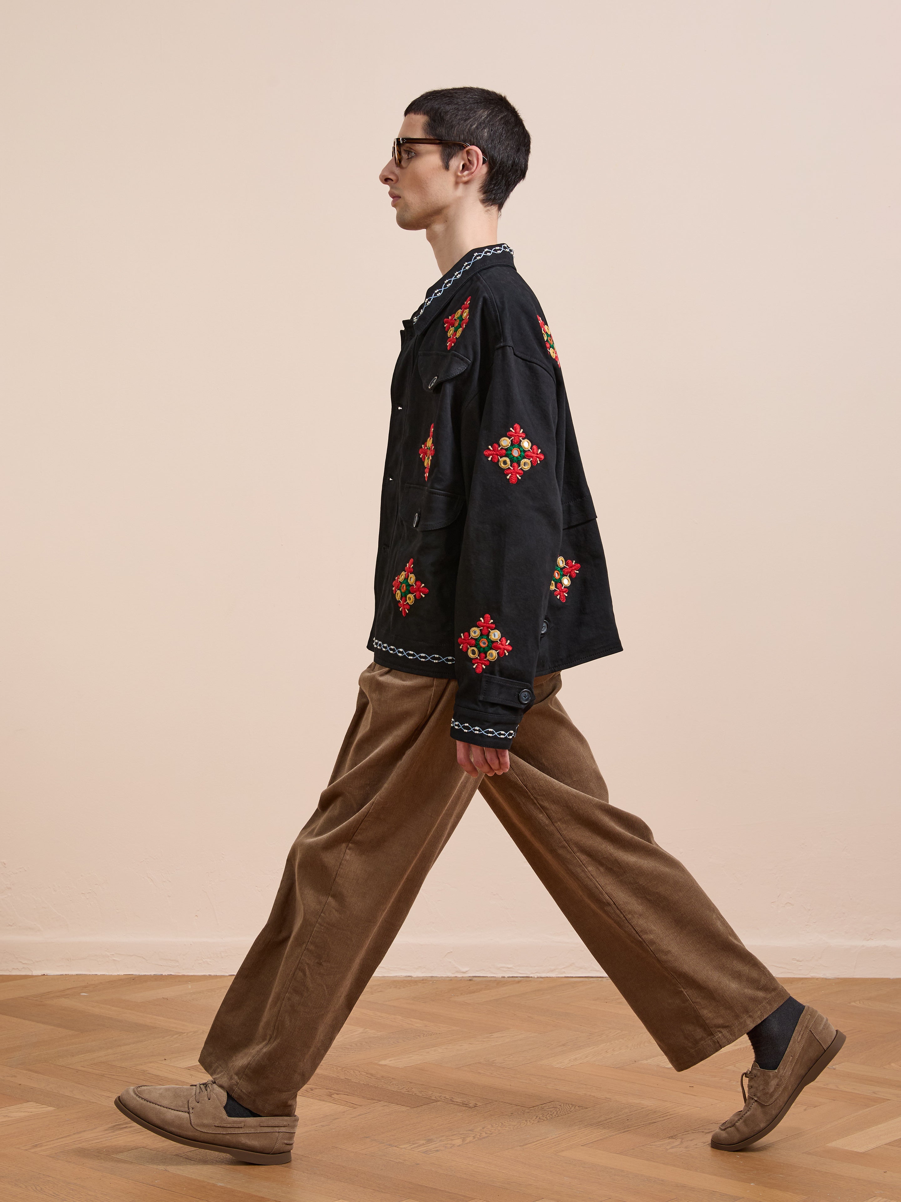 A person wearing glasses and the Found Mirror Ornament Hand Embroidered Work Jacket, featuring black fabric with red and green designs, plus brown pants and loafers, walks indoors on a wooden floor.
