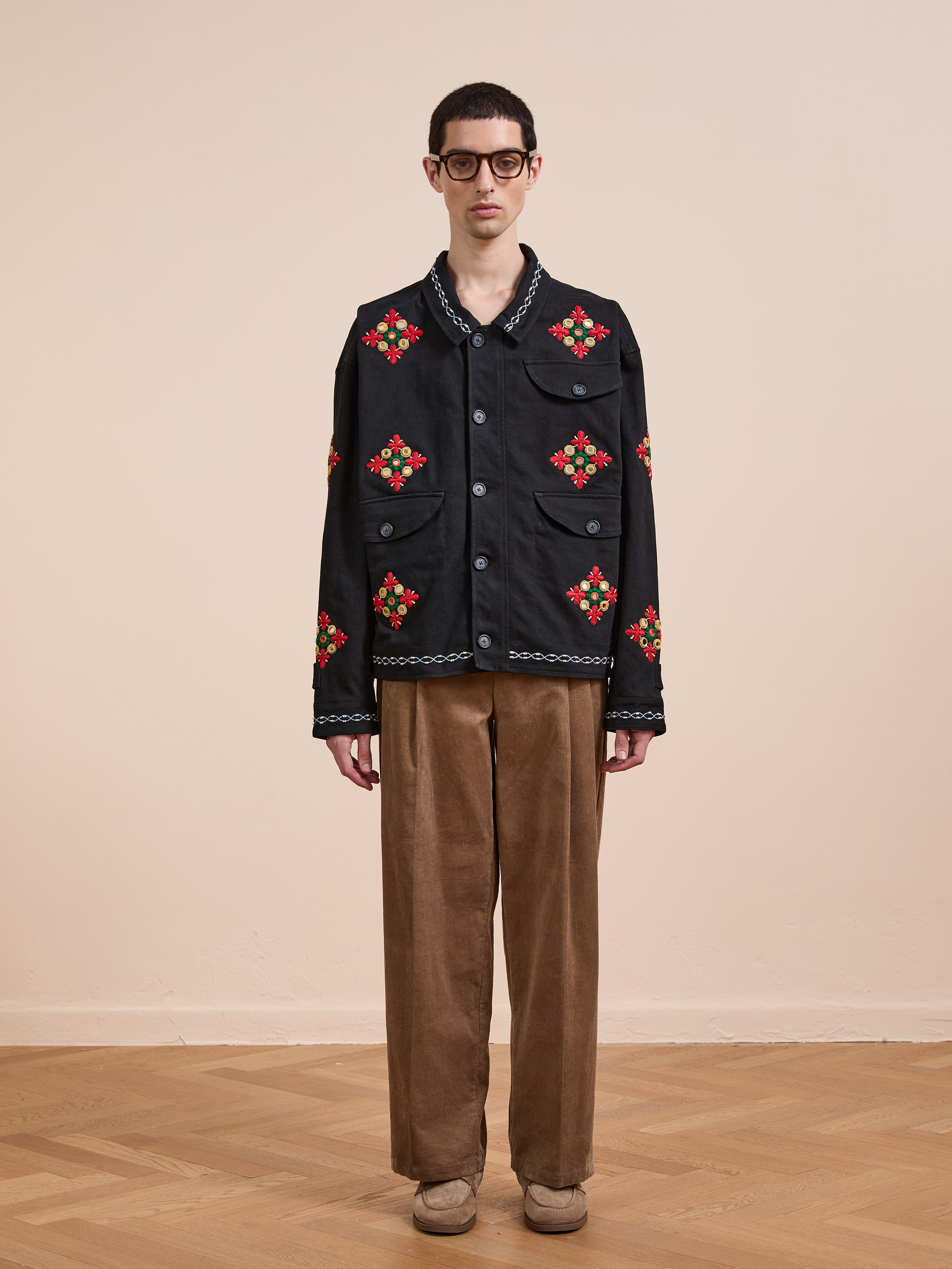A person stands indoors wearing glasses, the Found Mirror Ornament Hand Embroidered Work Jacket, brown corduroy pants, and beige shoes on a wooden floor with a plain beige background.
