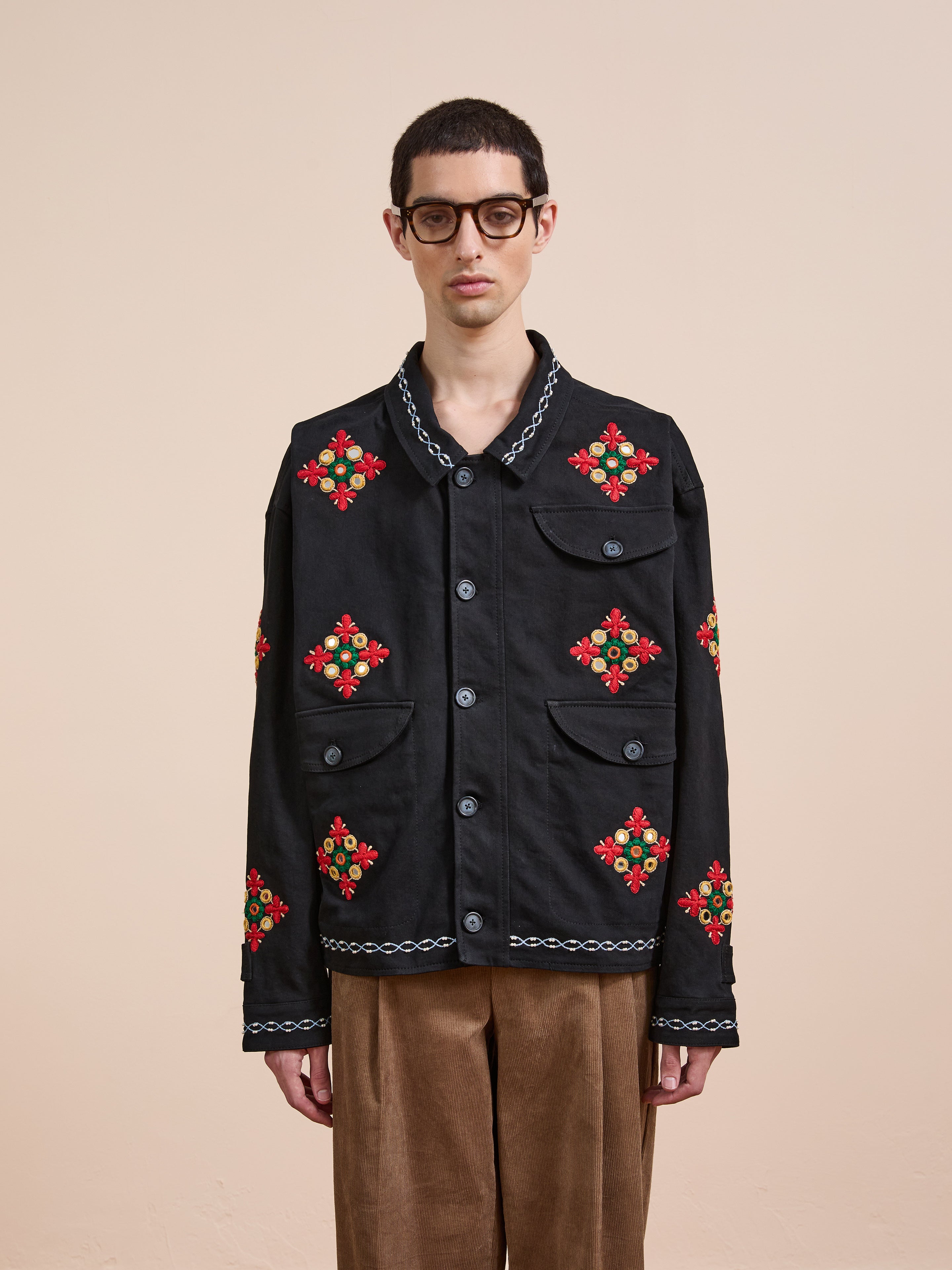 A person wearing glasses, brown pants, and the Found Mirror Ornament Hand Embroidered Work Jacket stands against a plain beige background.
