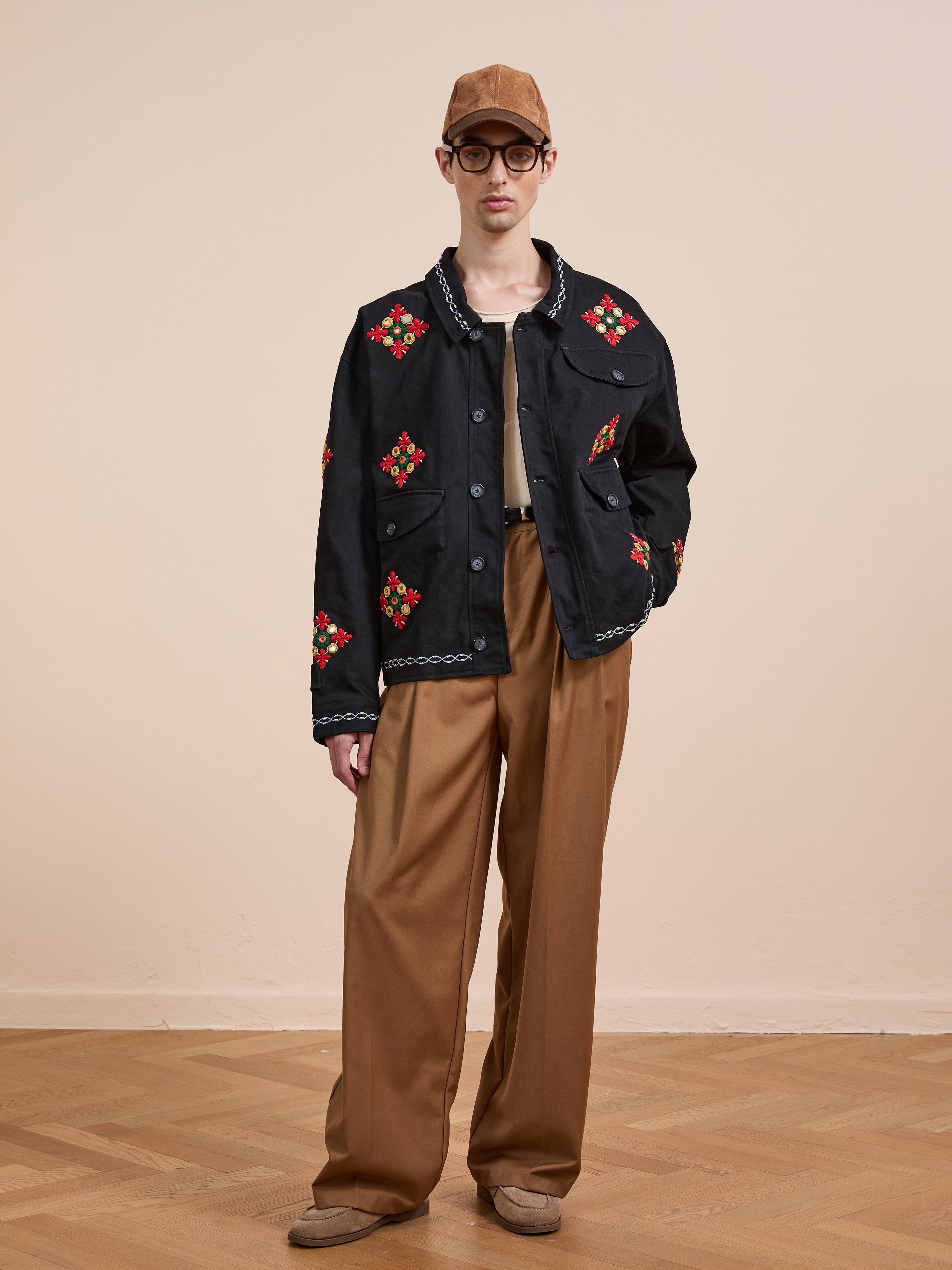 Indoors, a person wears Found’s Mirror Ornament Hand Embroidered Work Jacket in black, styled with a brown cap, round glasses, tan shirt, wide-leg brown pants, and brown shoes on a wooden floor.