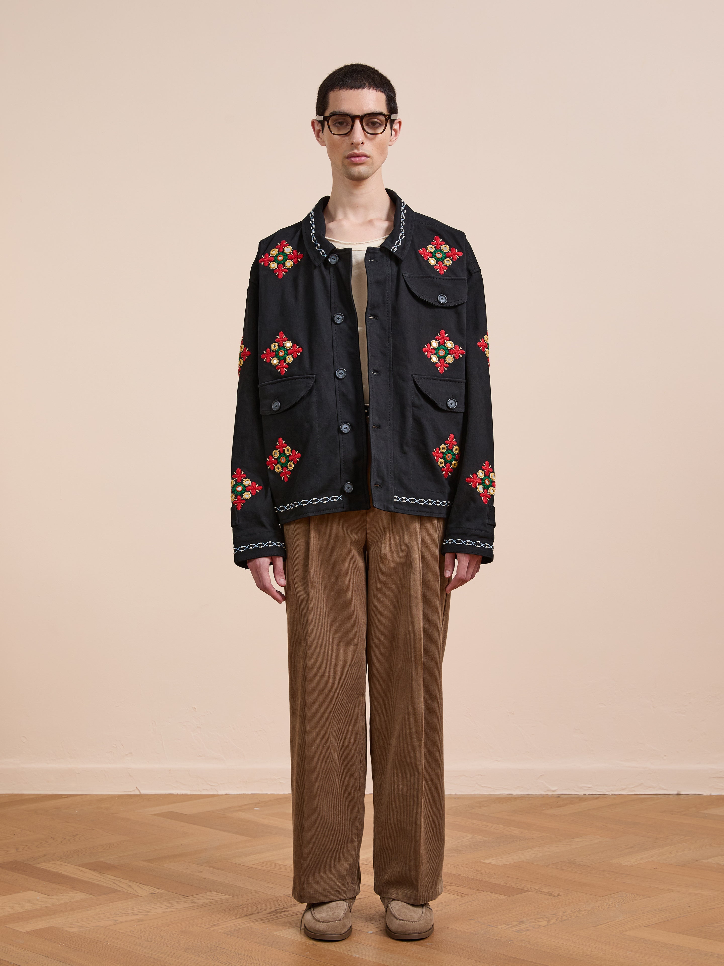 Indoors, a person wears glasses, a tan shirt, brown corduroy pants, beige shoes, and the Found Mirror Ornament Hand Embroidered Work Jacket featuring black fabric with red and green embroidery.