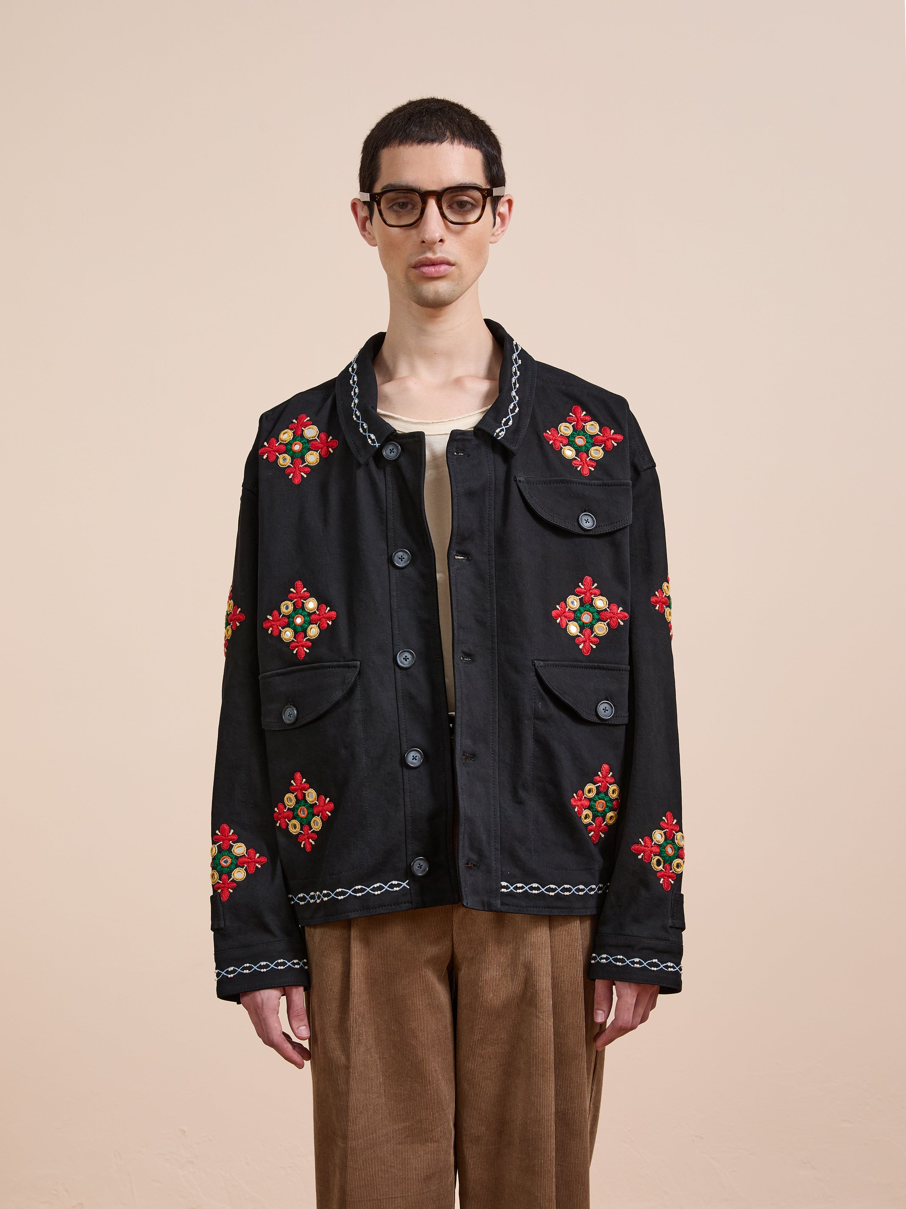 A person wearing glasses and brown pants models the Found Mirror Ornament Hand Embroidered Work Jacket against a plain beige background.
