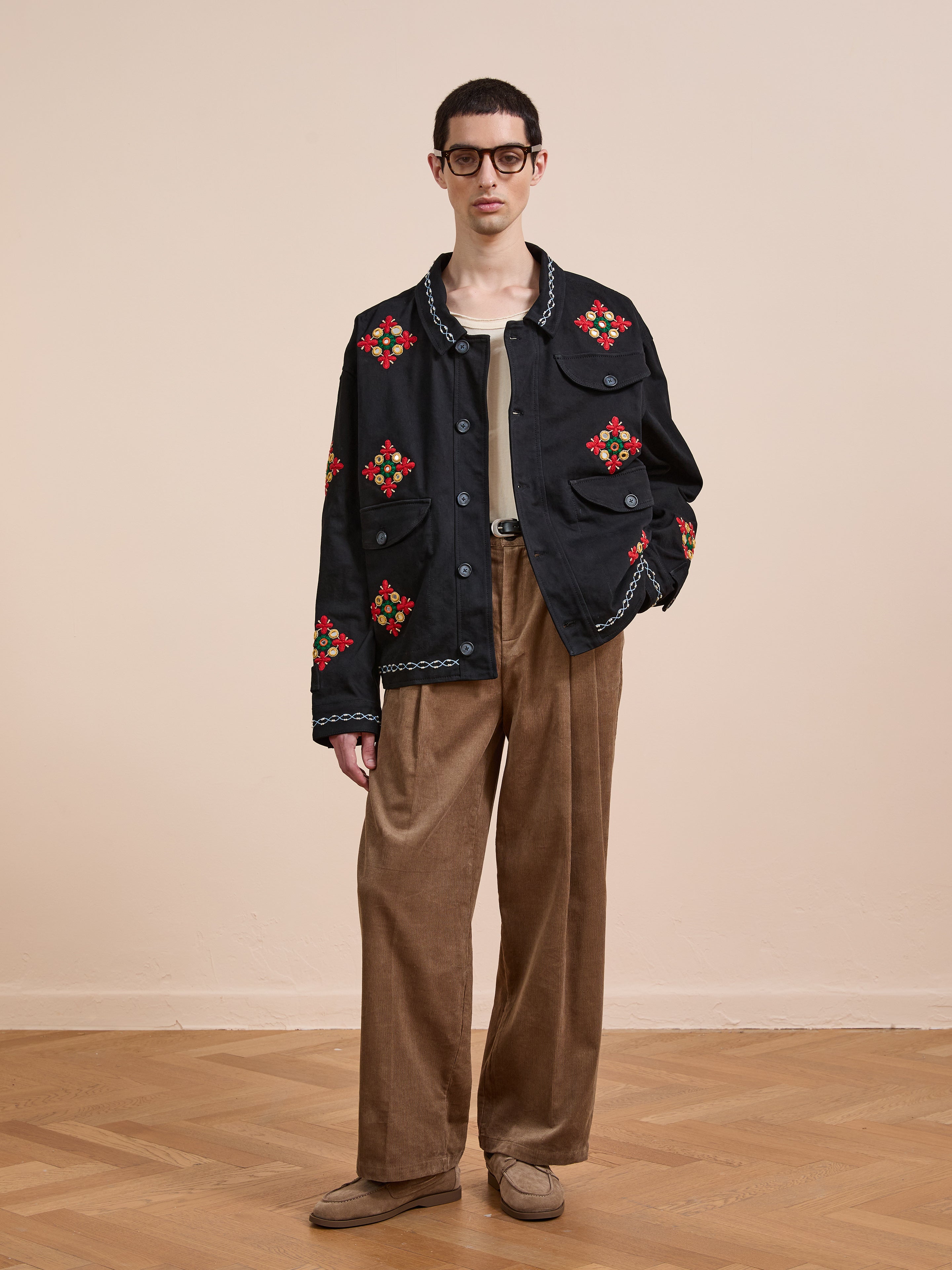 Indoors, a person wears the Found Mirror Ornament Hand Embroidered Work Jacket in black, paired with a tan shirt, brown wide-leg corduroy pants, brown shoes, and glasses.