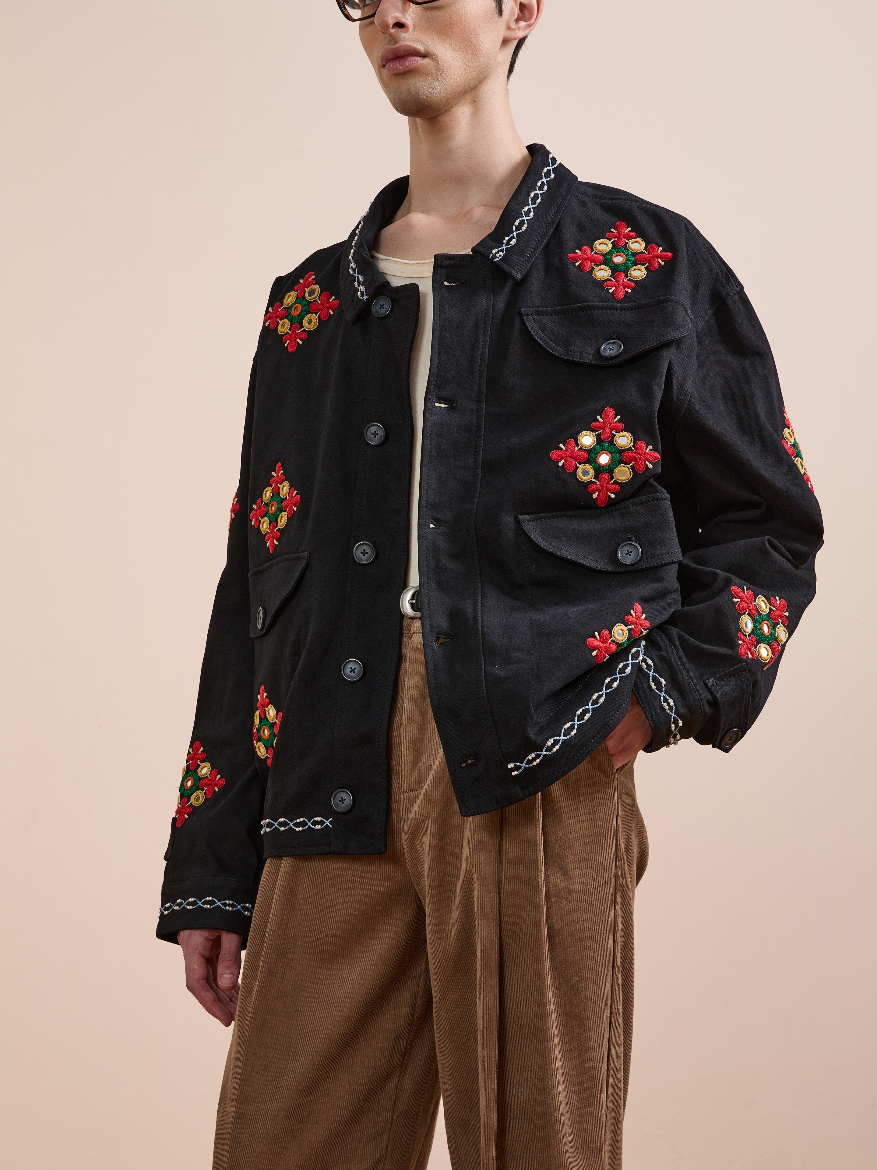 A person wearing Found’s Mirror Ornament Hand Embroidered Work Jacket in black with red and green floral details, paired with a beige shirt and brown pleated pants, stands against a plain light background.