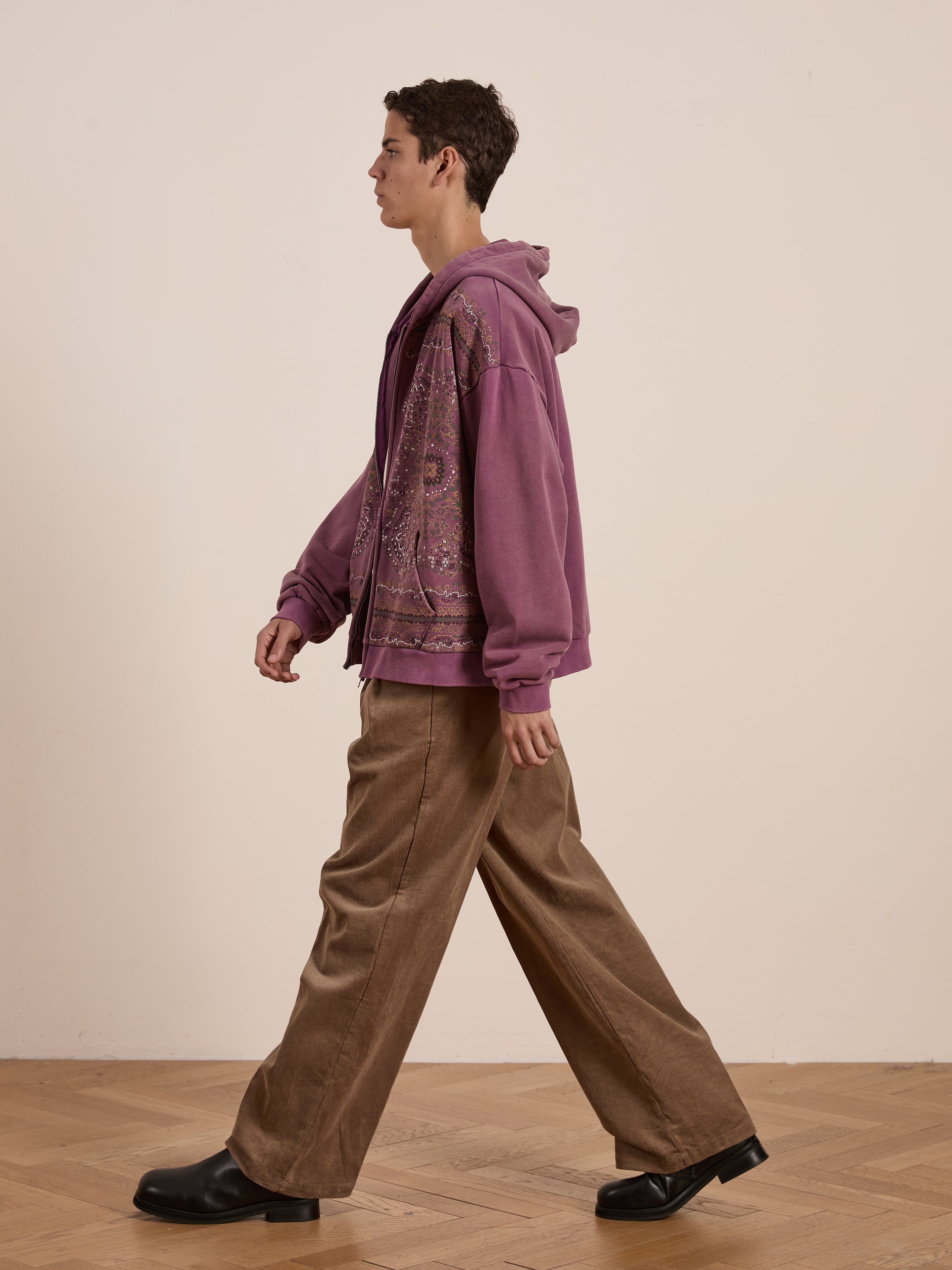 A person walks sideways on a wooden floor wearing FOUND's Motif Zip-Up Hoodie in mauve, paired with brown wide-leg pants and black shoes—a streetwear essential.