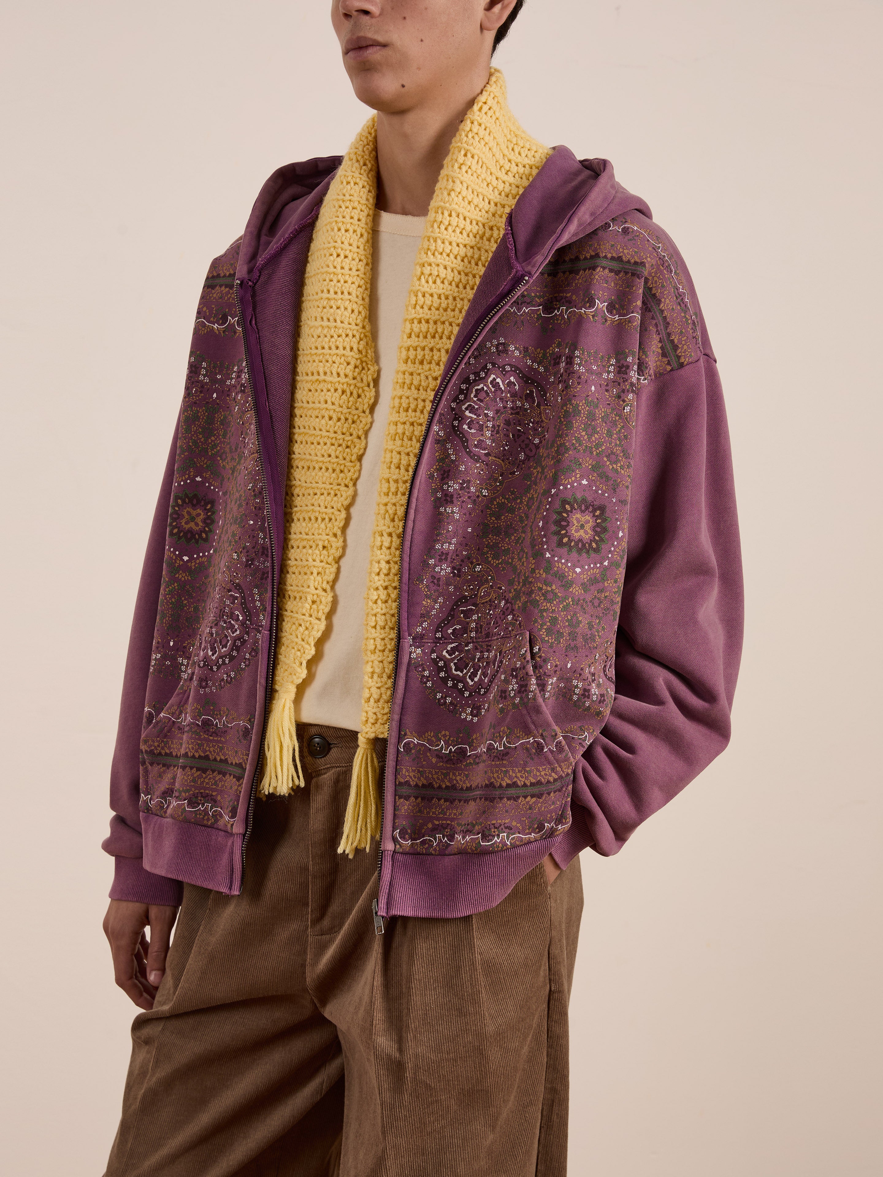 Wearing the FOUND Motif Zip-Up Hoodie, adorned in its purple pattern, a person completes their streetwear look with a yellow scarf and brown pants against a neutral backdrop.