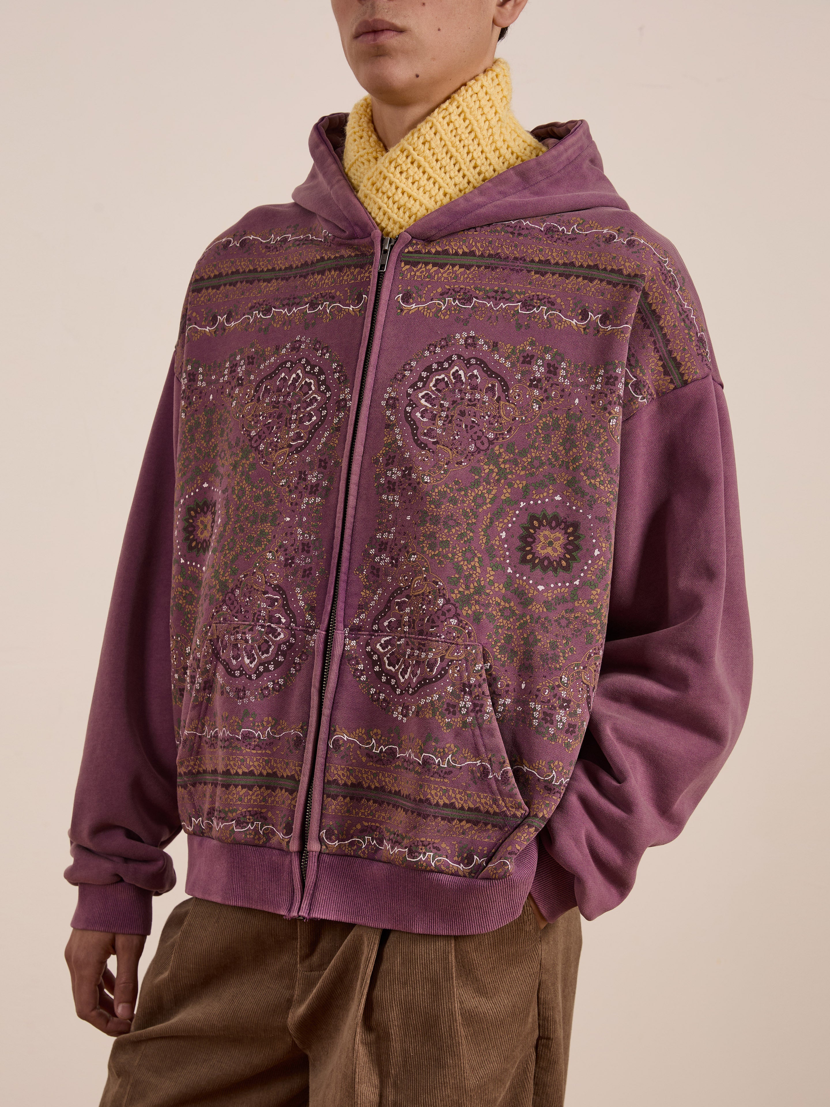 A person stands against a plain backdrop, wearing FOUND's Motif Zip-Up Hoodie in purple pattern over a yellow knit scarf and brown pants, showcasing streetwear essentials.