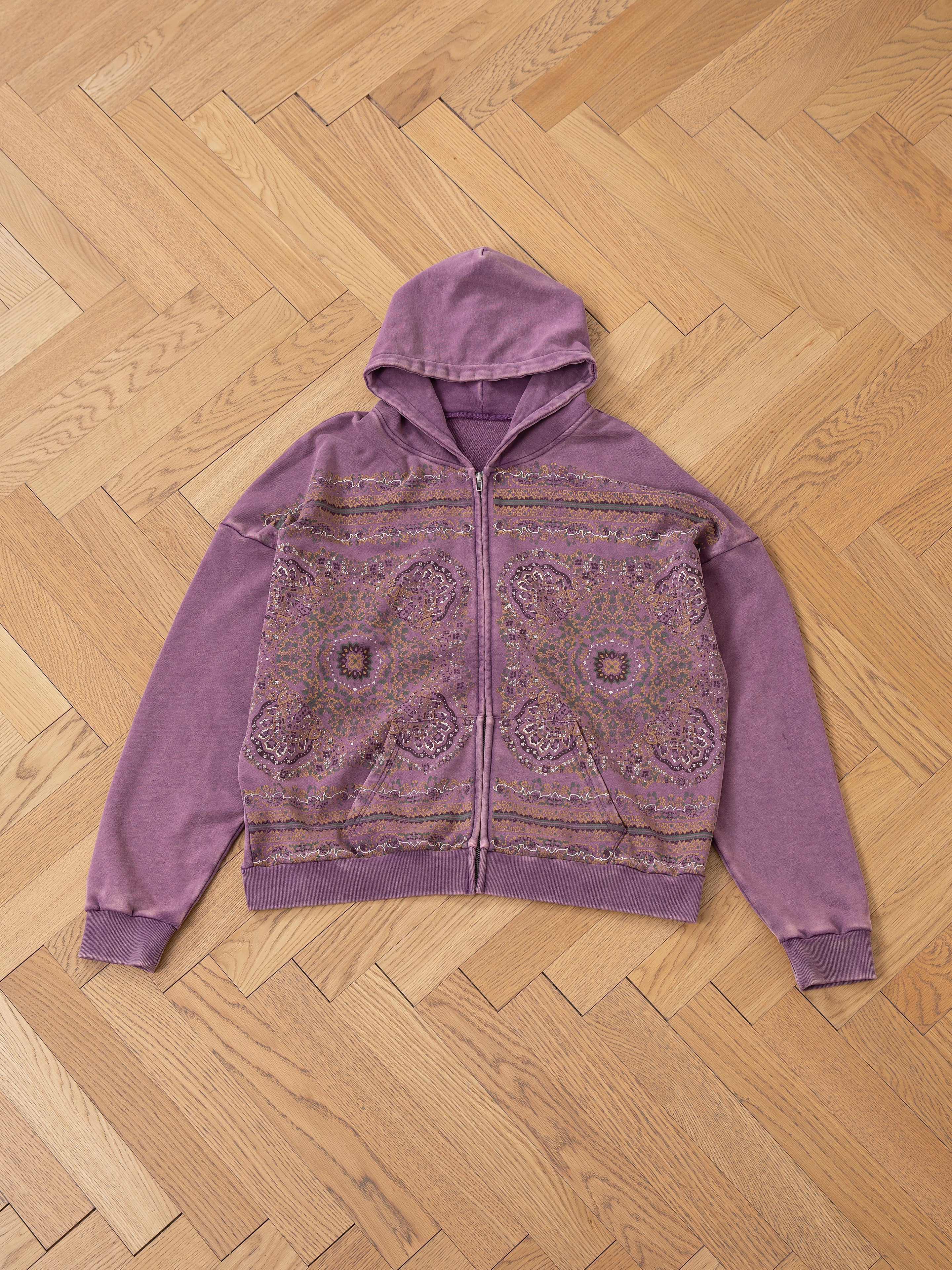 The Motif Zip-Up Hoodie by FOUND, showcasing intricate mandala art in purple, lies flat on a wooden herringbone floor—a streetwear essential.