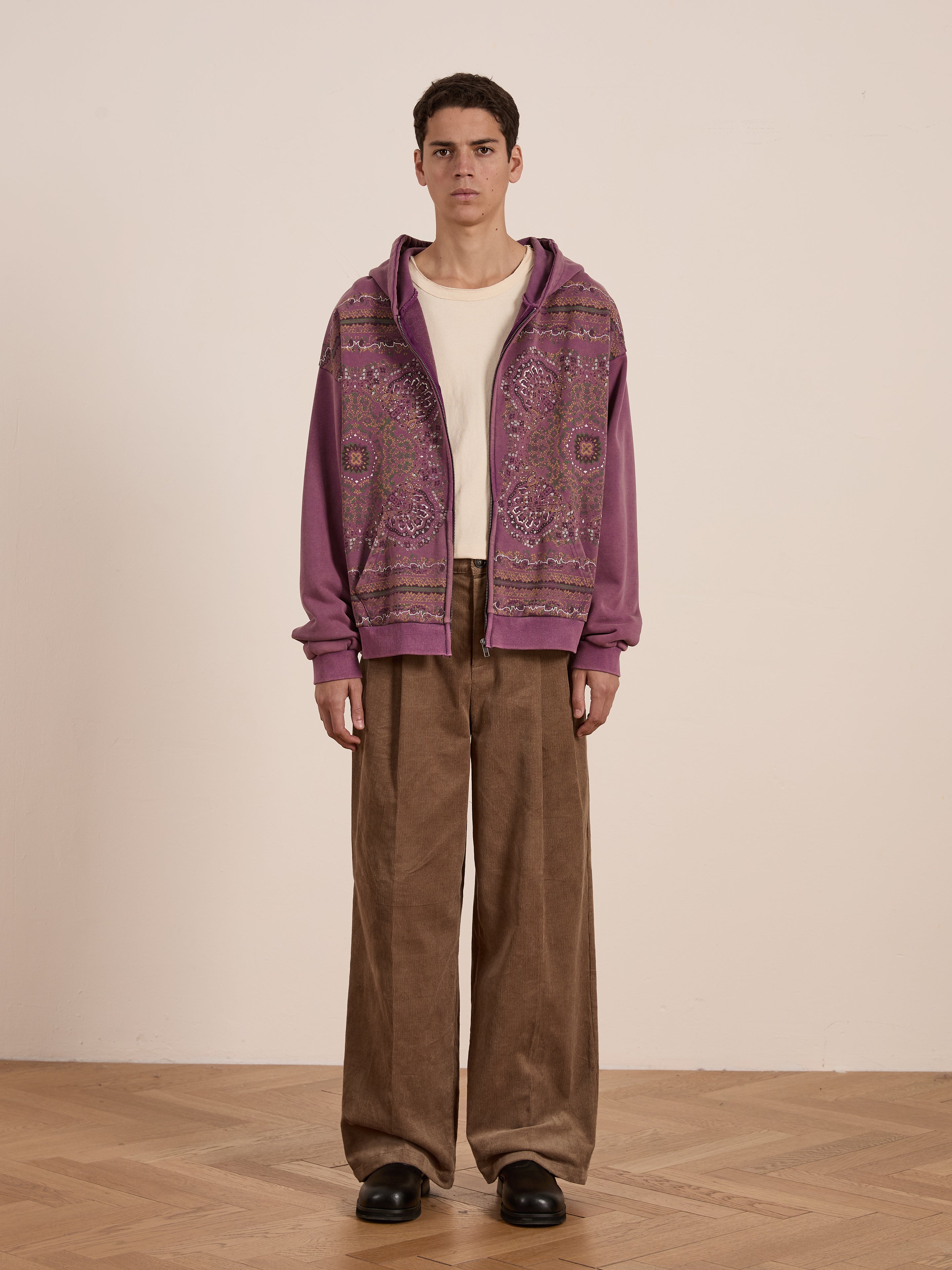 Dressed in FOUND's Motif Zip-Up Hoodie featuring purple mandala patterns, a person pairs it with a white shirt and brown wide-legged pants, standing gracefully on a wooden floor against a plain backdrop.