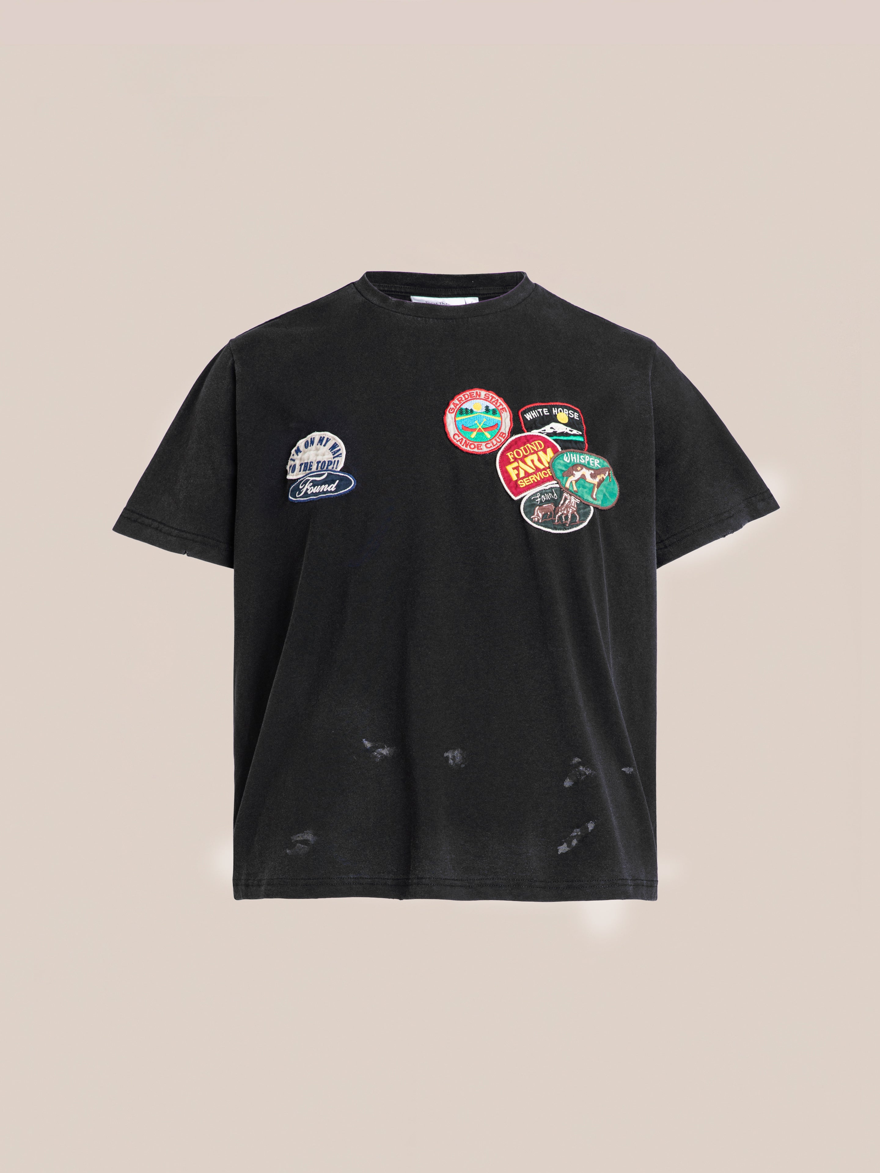 FOUND's Multi Patch Washed Tee in black, crafted from enzyme-washed cotton, showcases multiple colorful patches on the front and is presented against a plain beige background.