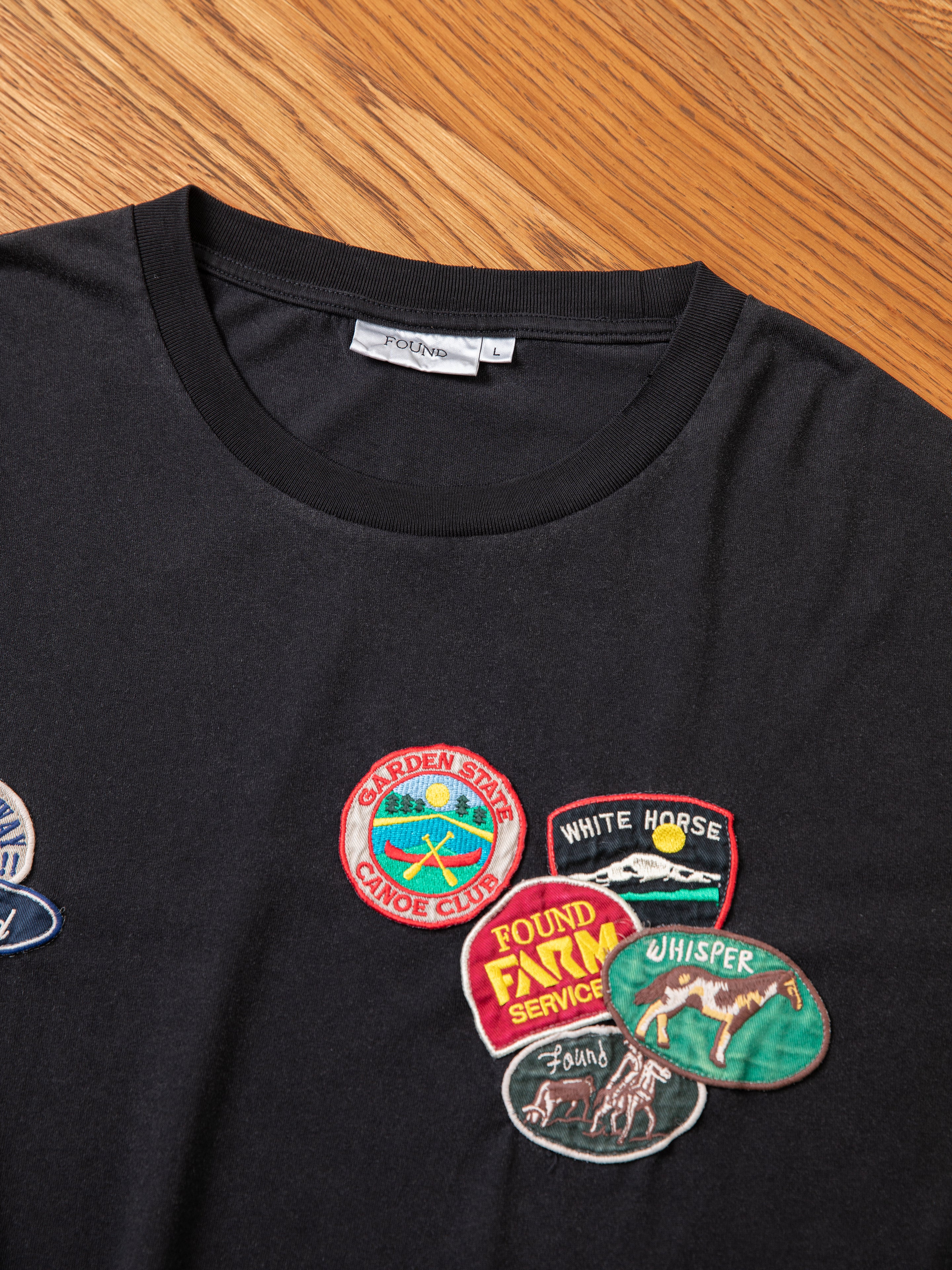 The Multi Patch Washed Tee by FOUND is a black t-shirt featuring embroidered patches such as "GARDEN STATE CANOE CLUB," "WHITE HORSE," "FOUND FILM SERVICE," and "WILDERNESS: Whisper: AWARD." The shirt is displayed on a wooden surface.