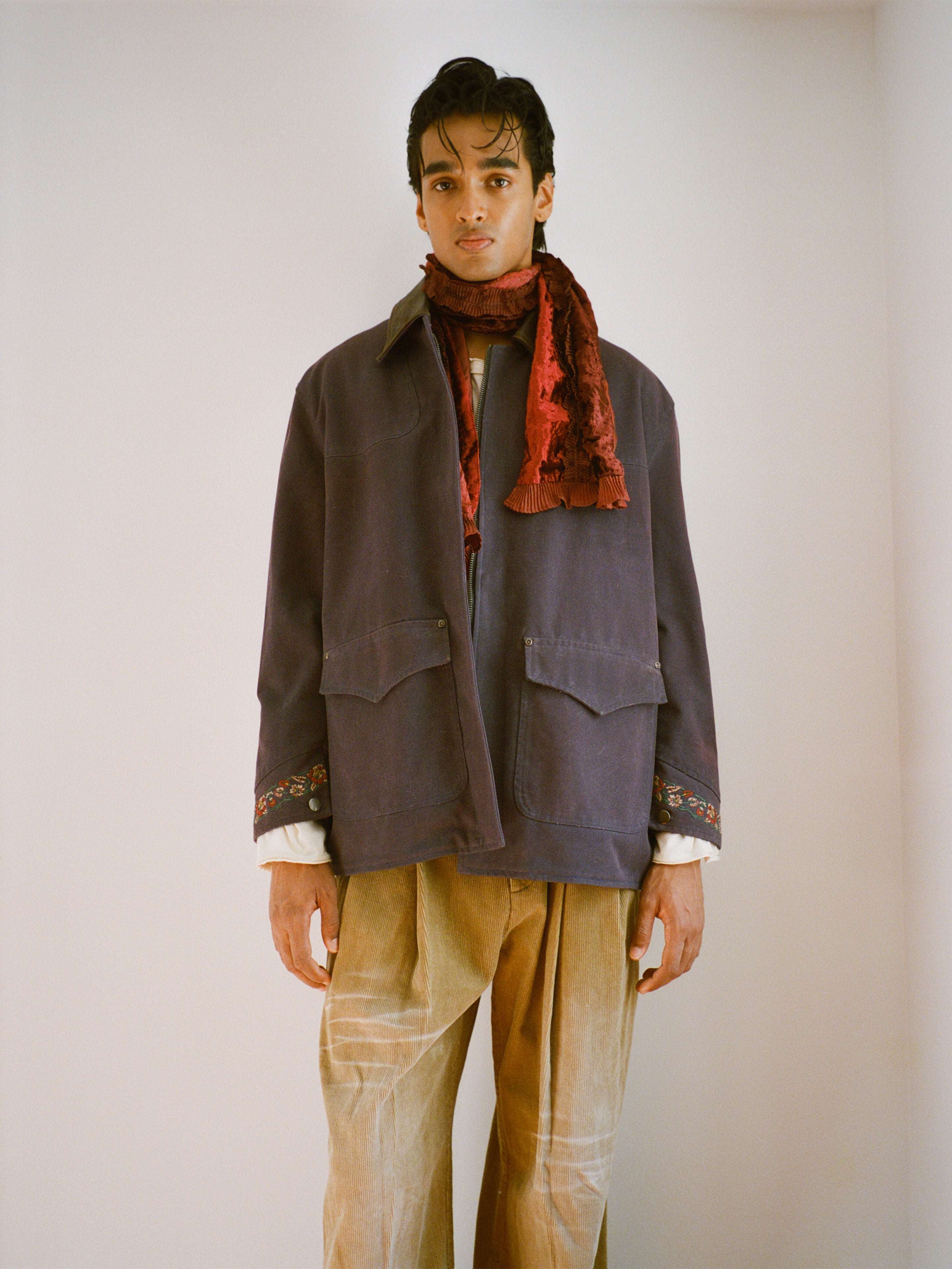 A person stands indoors facing the camera against a plain background, wearing the Found Canvas Distressed Barn Jacket in navy with a distressed leather collar, styled in a unisex relaxed fit with light pants, a cream shirt, and a textured red scarf.