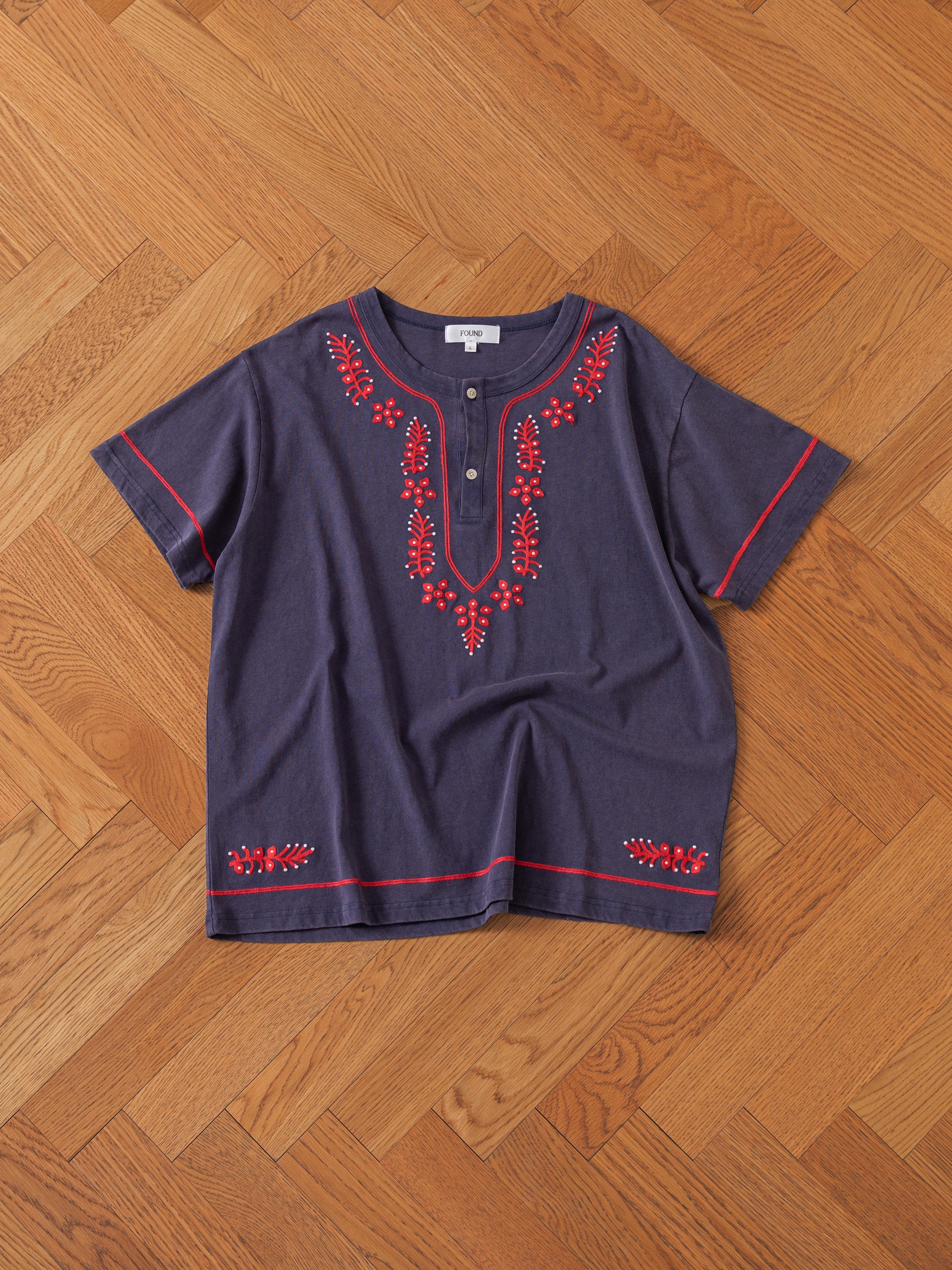 A FOUND Embroidered Henley, featuring blue fabric with red embroidery on the front and sleeves along with a buttoned neckline, laid flat on a wooden floor with a herringbone pattern.