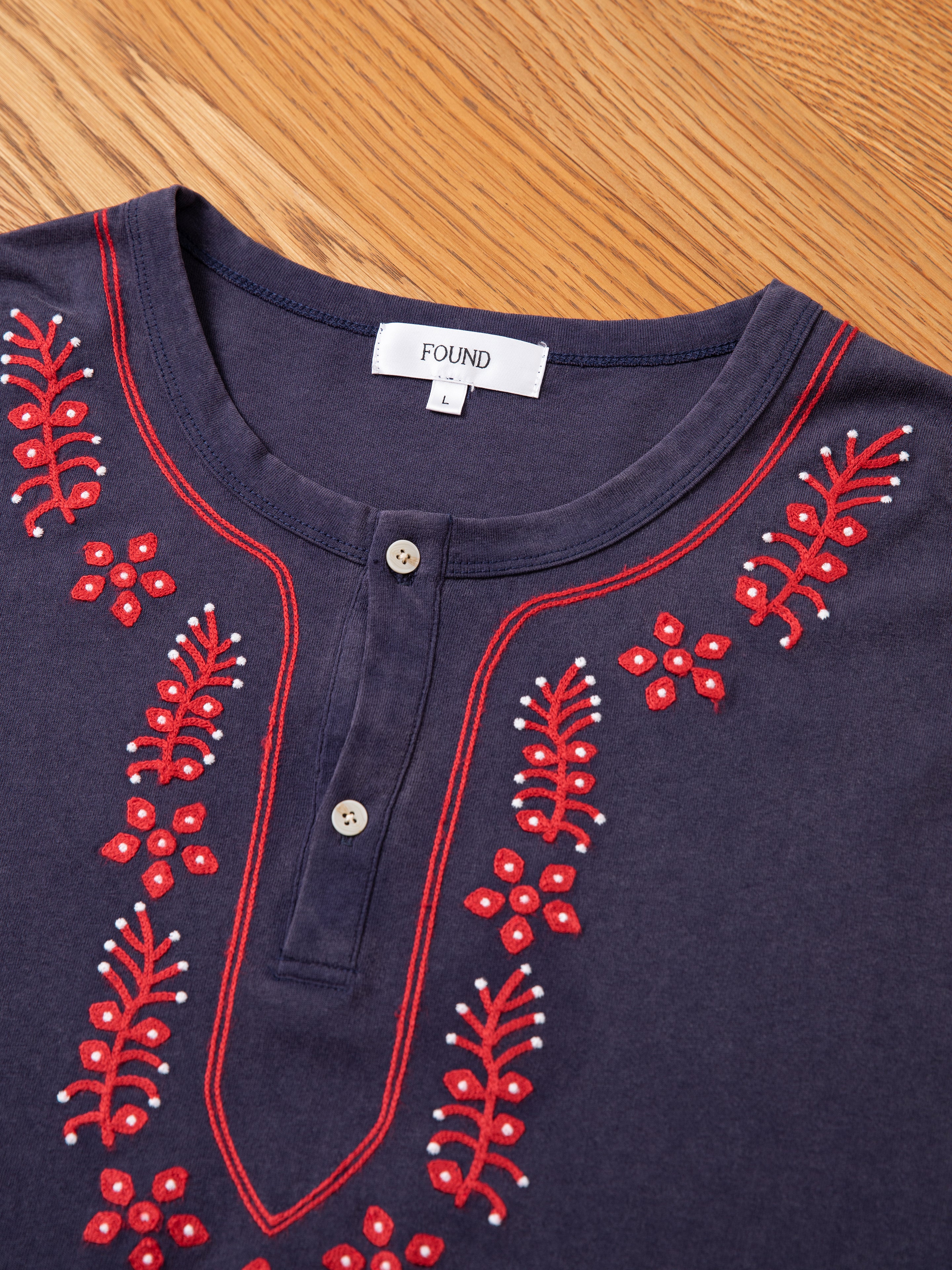 The Embroidered Henley by FOUND features a dark blue color with red and white floral embroidery near the neckline, and is photographed on a wooden surface.