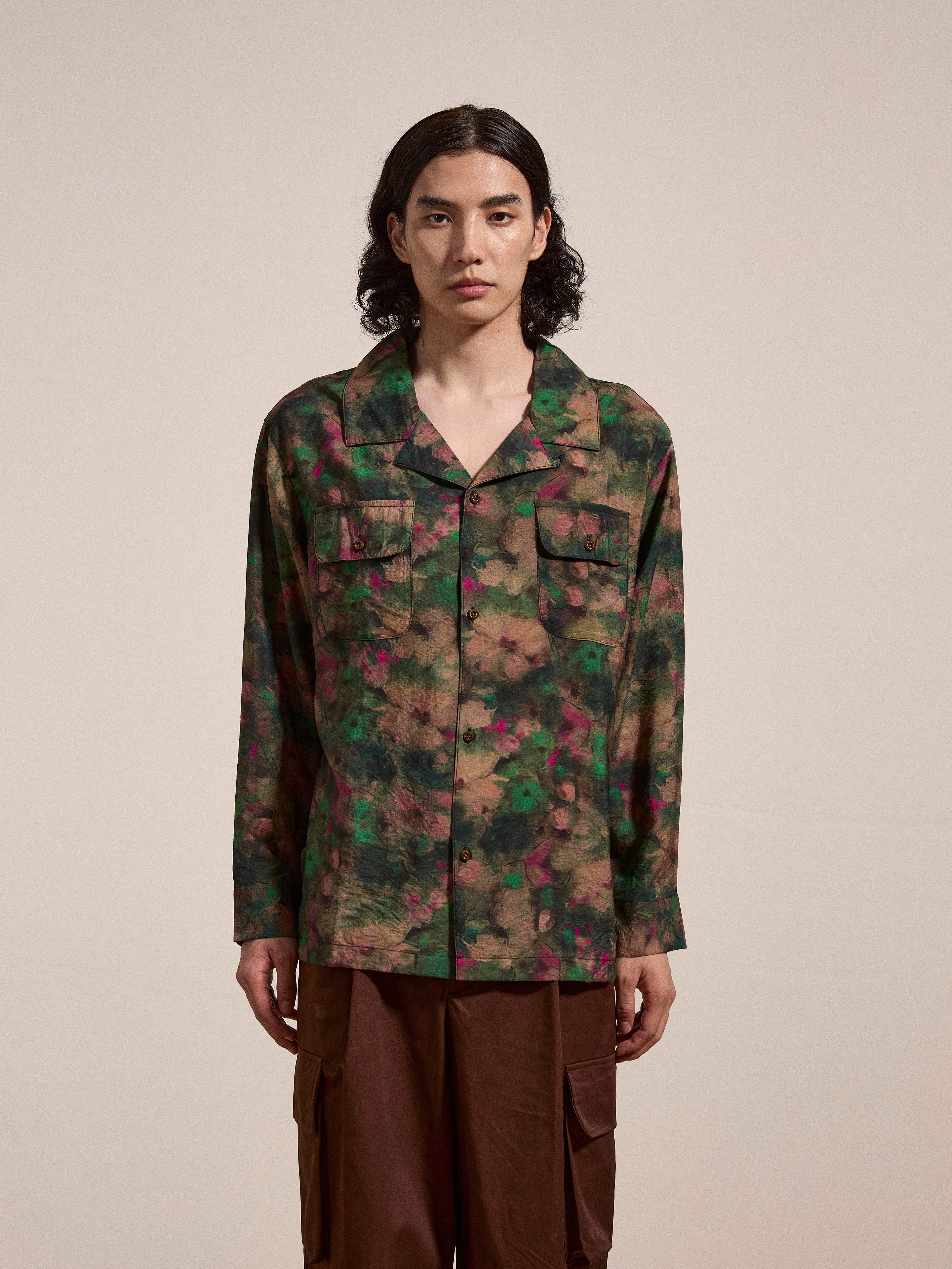 A person with long hair is standing against a plain background, wearing a green and red patterned Charlot LS Camp Shirt from FOUND, featuring dual chest pockets, paired with brown pants.