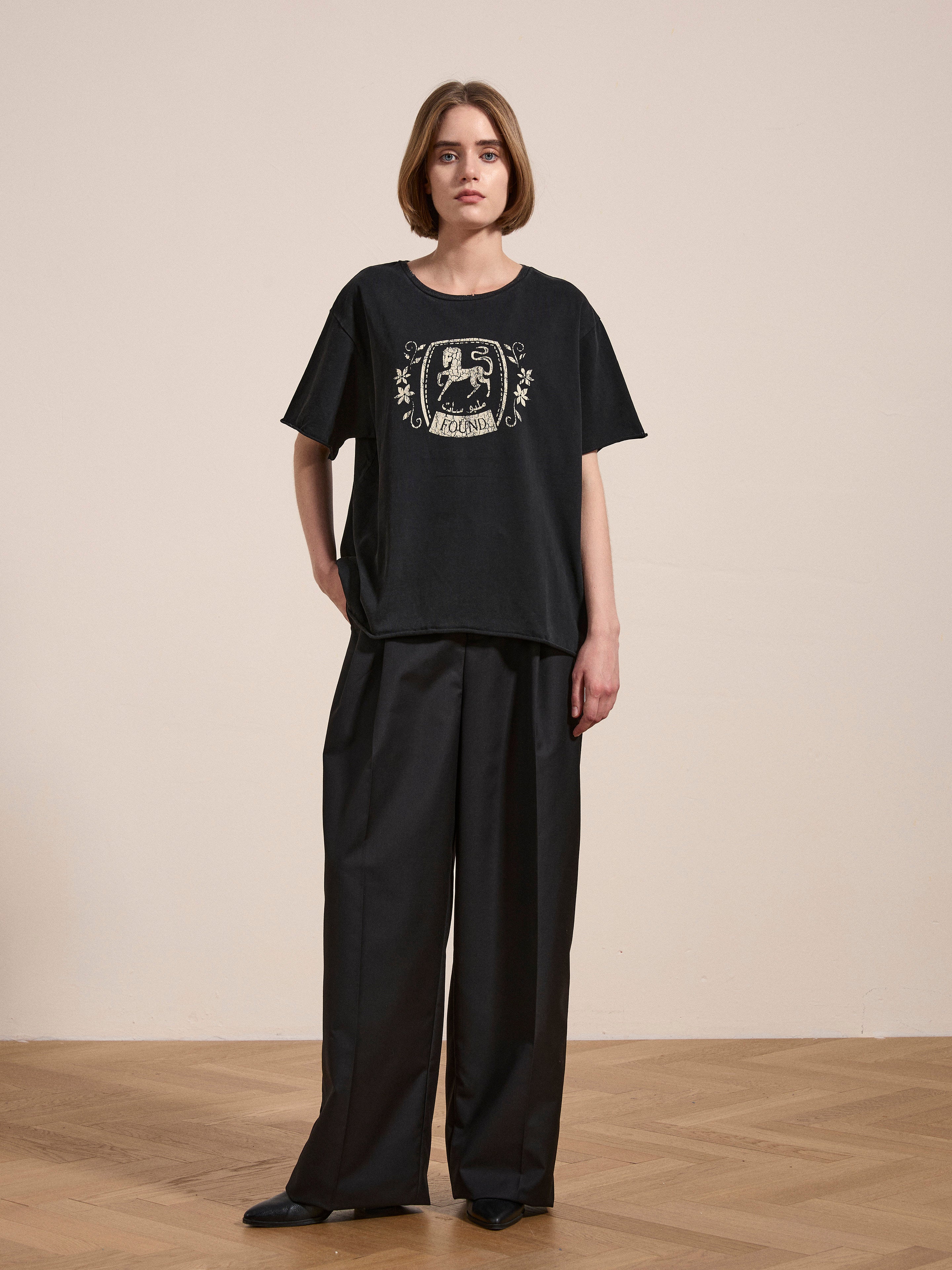 A person stands on a wooden floor wearing a Horse Logo Print Tee by FOUND, made from 100% cotton with an enzyme wash, paired with loose-fitting black pants.