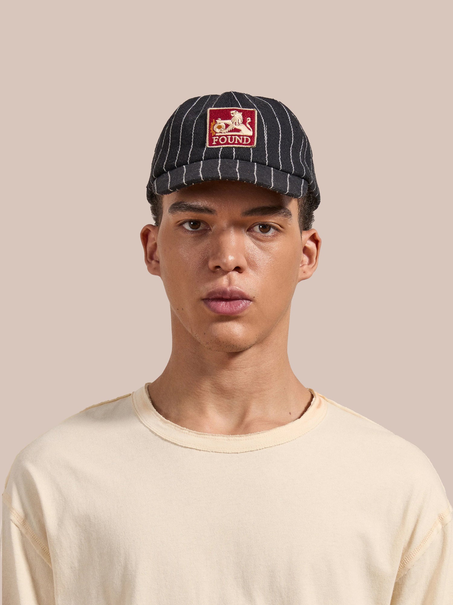 Person wearing a Pinstripe Lion Cap from FOUND, paired with a beige shirt, stands against a plain beige background.