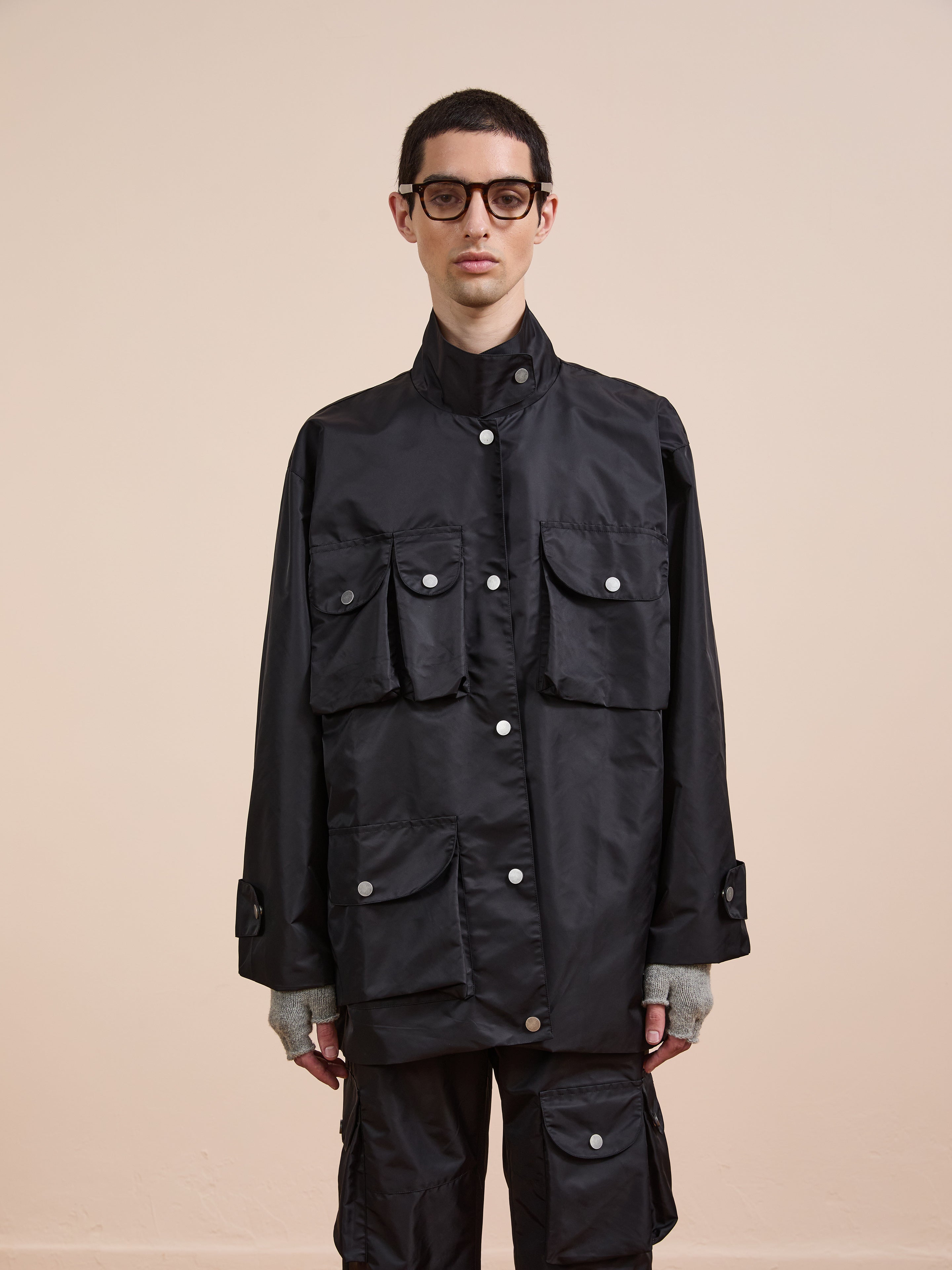 A man wearing glasses and a black FOUND Cargo Nylon Jacket with multiple front pockets stands against a plain beige background.