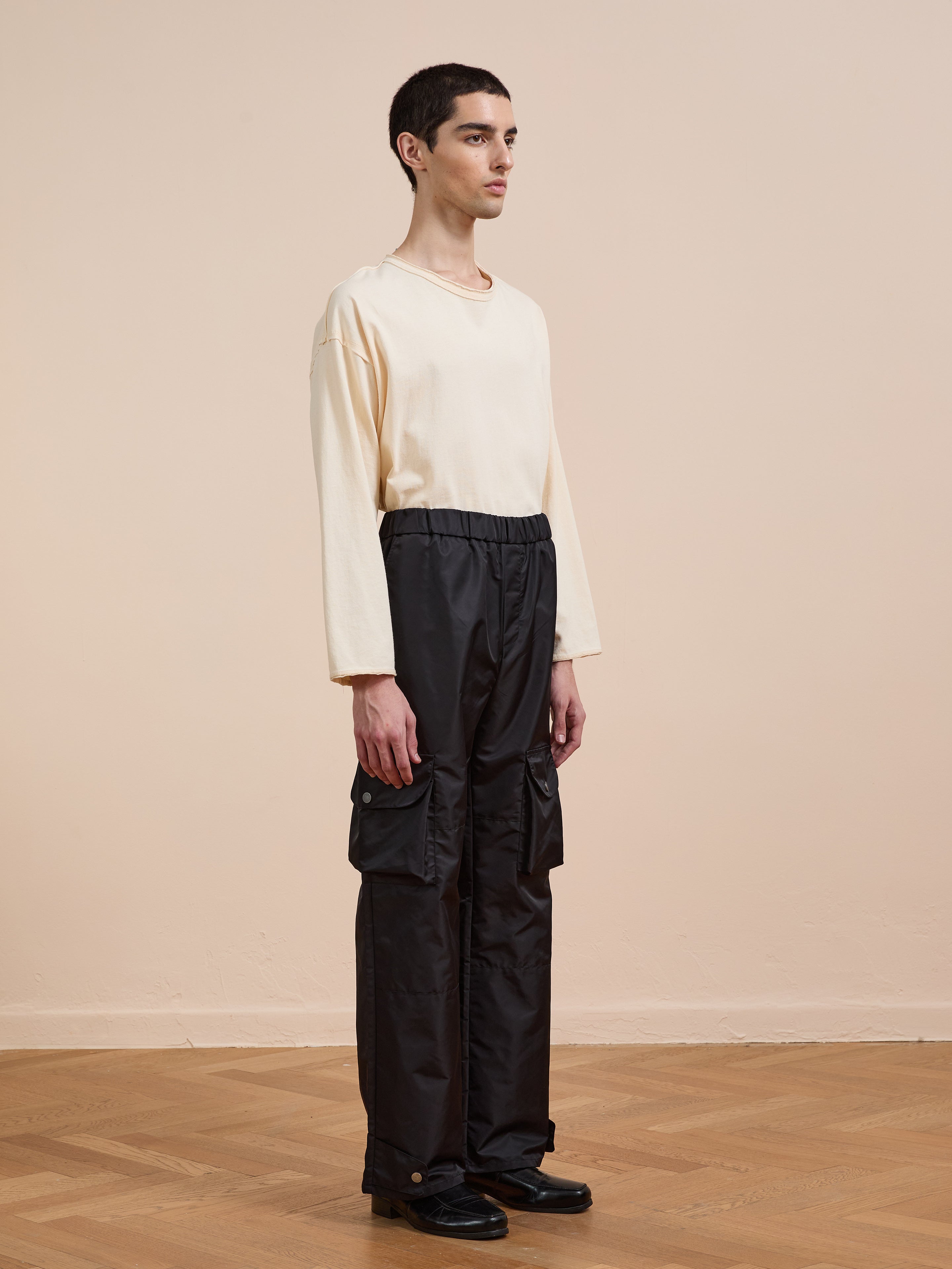 A person stands indoors in a relaxed pose wearing FOUND Nylon Cargo Pants with large side pockets and a cream long-sleeve shirt, facing slightly to the side.
