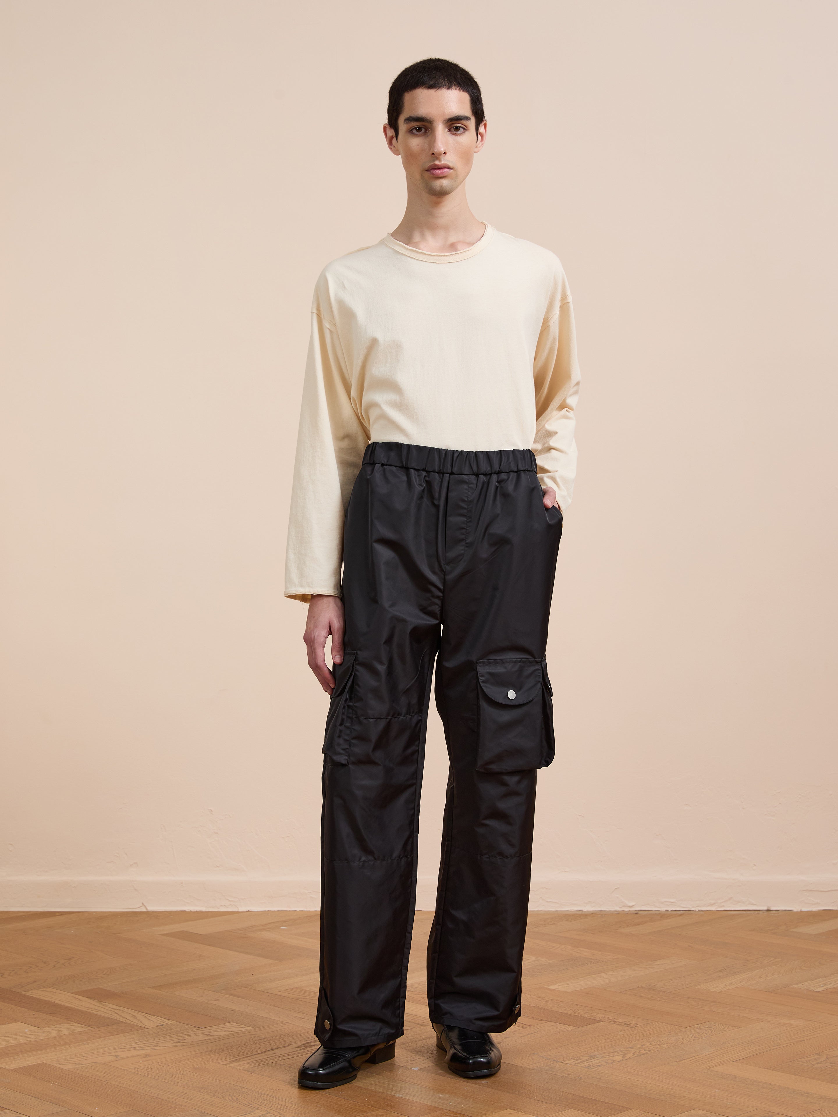 A person stands indoors on a wooden floor against a plain beige wall, wearing FOUND Nylon Cargo Pants in black with pockets and a relaxed-fit beige long-sleeve shirt.