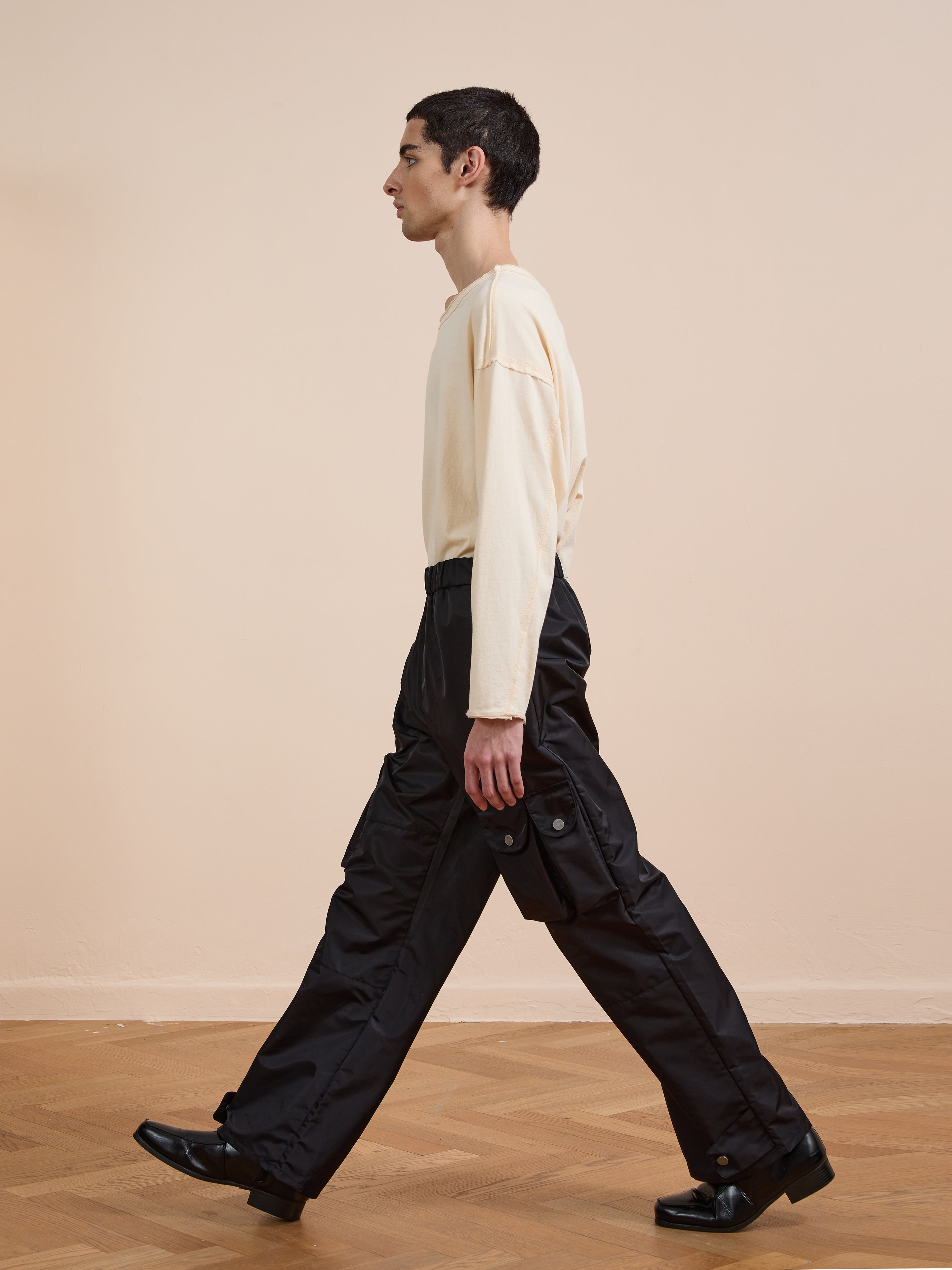 A person with short dark hair walks indoors by a beige wall and wooden floor, wearing a cream long-sleeve shirt, black shoes, and relaxed fit FOUND Nylon Cargo Pants.