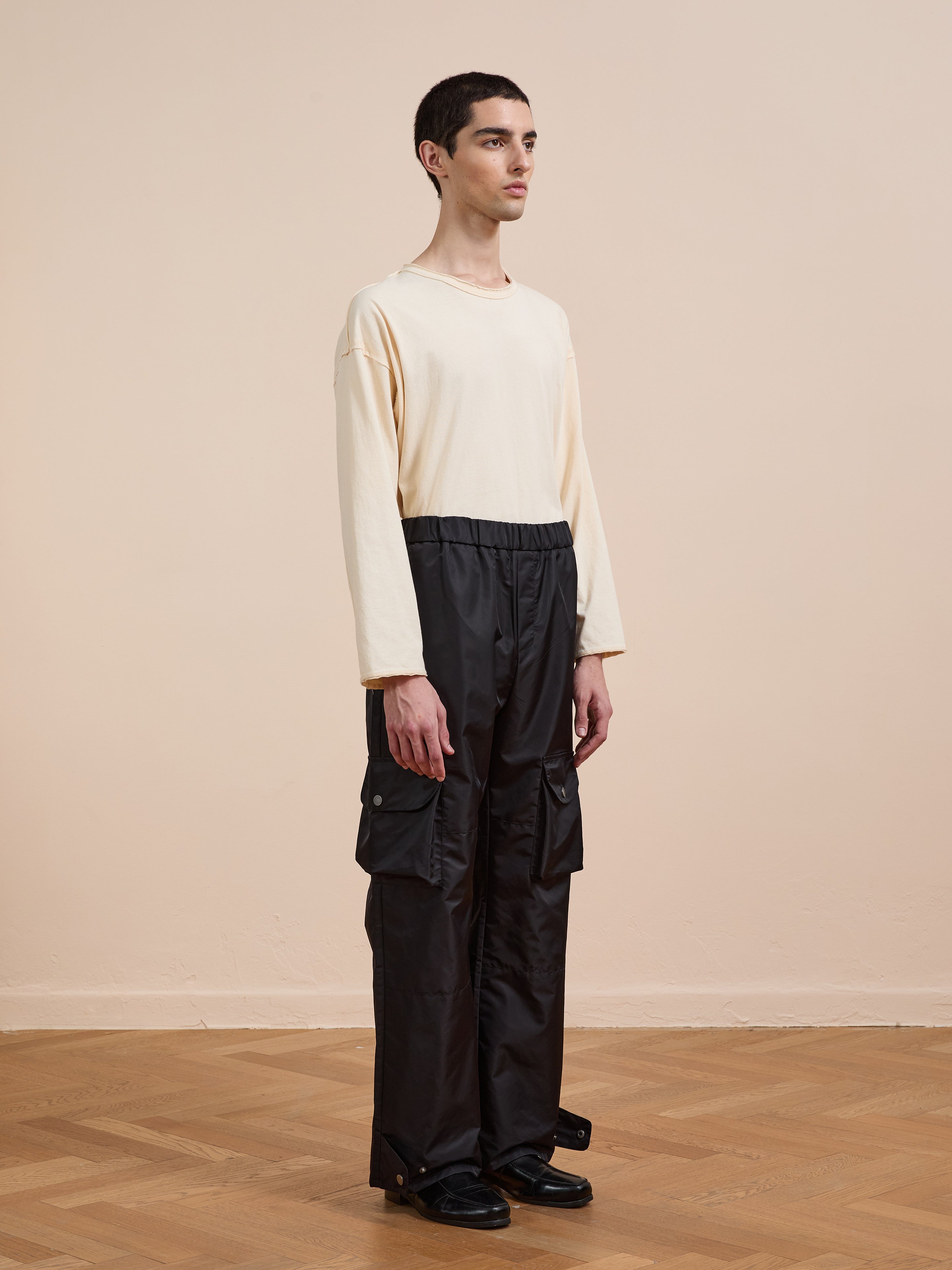 A person stands indoors on a wooden floor, wearing a relaxed beige long-sleeve shirt and FOUND Nylon Cargo Pants in black with black shoes, facing slightly to the right.
