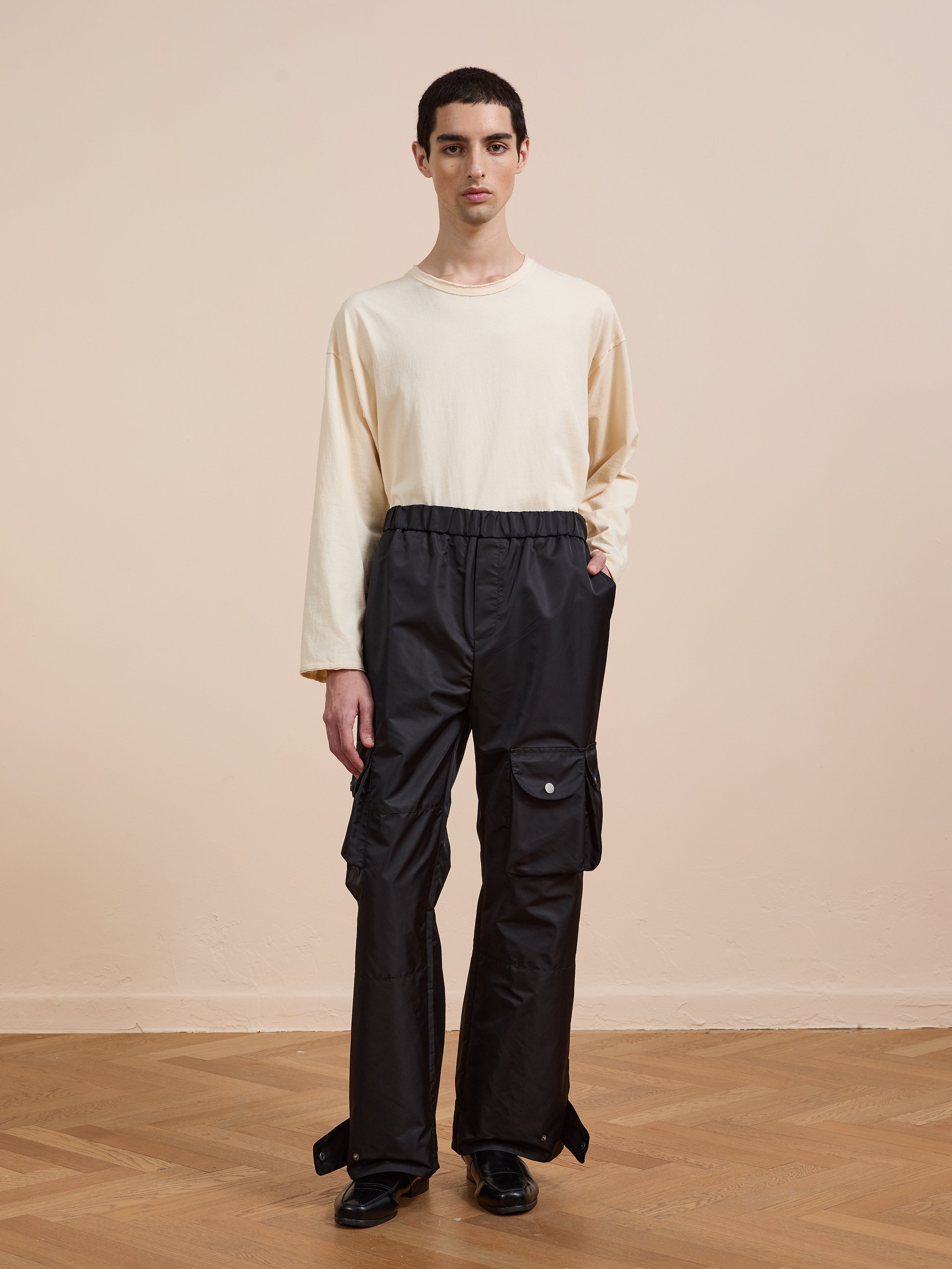 Indoors against a plain beige background, a person wears FOUND Nylon Cargo Pants in black with pockets, paired with a long-sleeve cream shirt and black shoes.