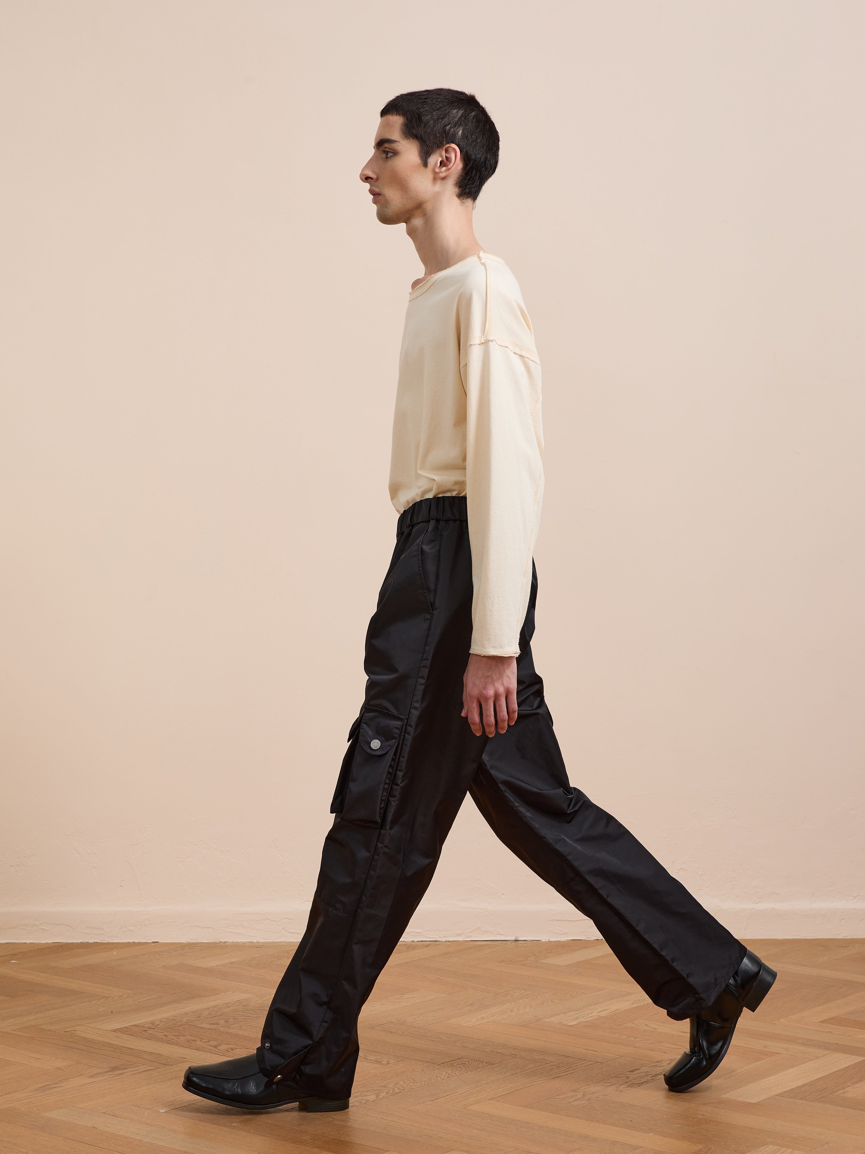 Wearing FOUND Nylon Cargo Pants in black, a person pairs them with a cream long-sleeve shirt and black shoes, walking indoors on a wooden floor against a beige wall.