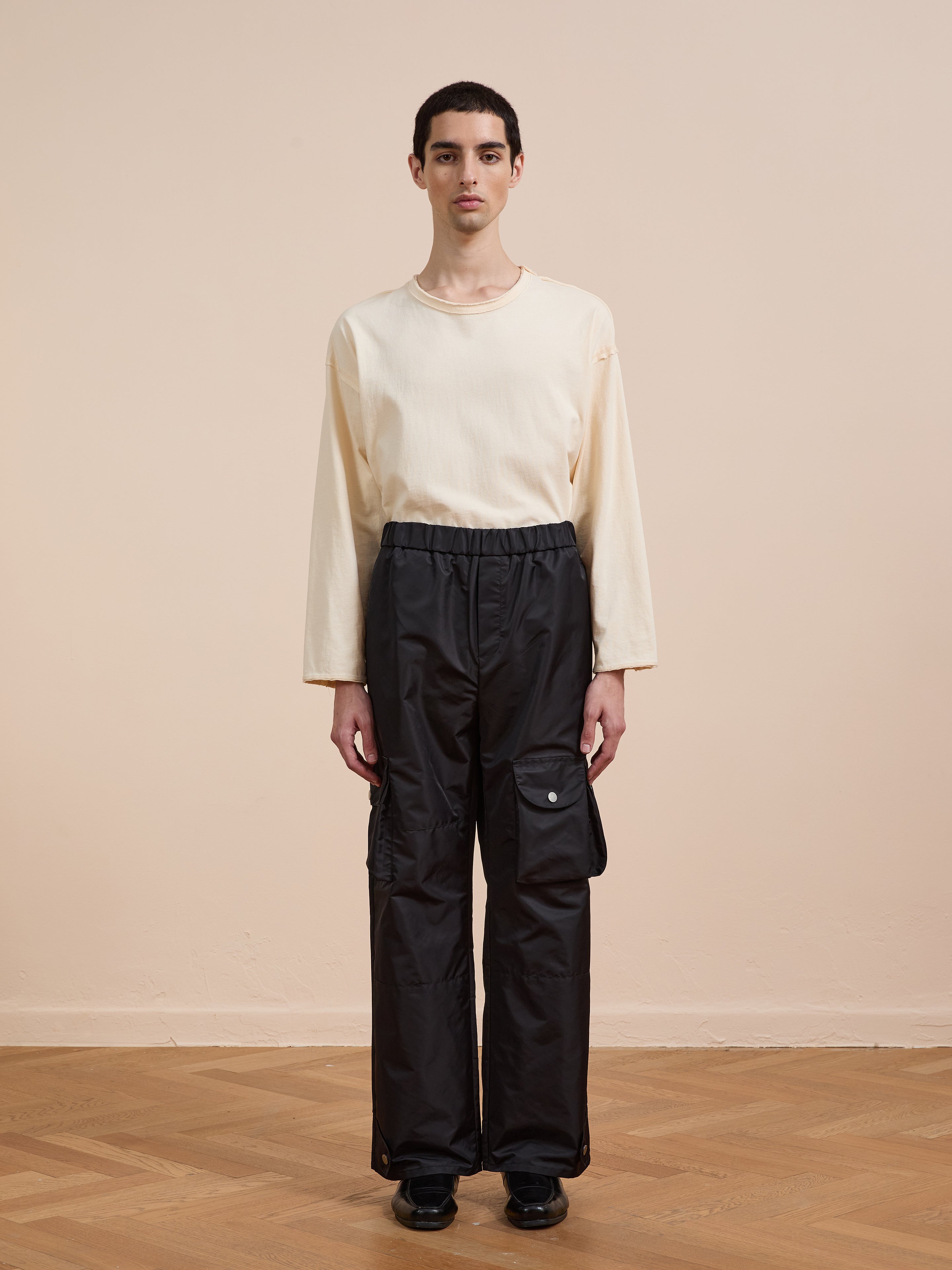 A person stands indoors on a wooden floor, wearing FOUND's black Nylon Cargo Pants—unisex with side pockets and a relaxed fit—paired with a cream long-sleeve shirt, facing the camera against a beige wall.