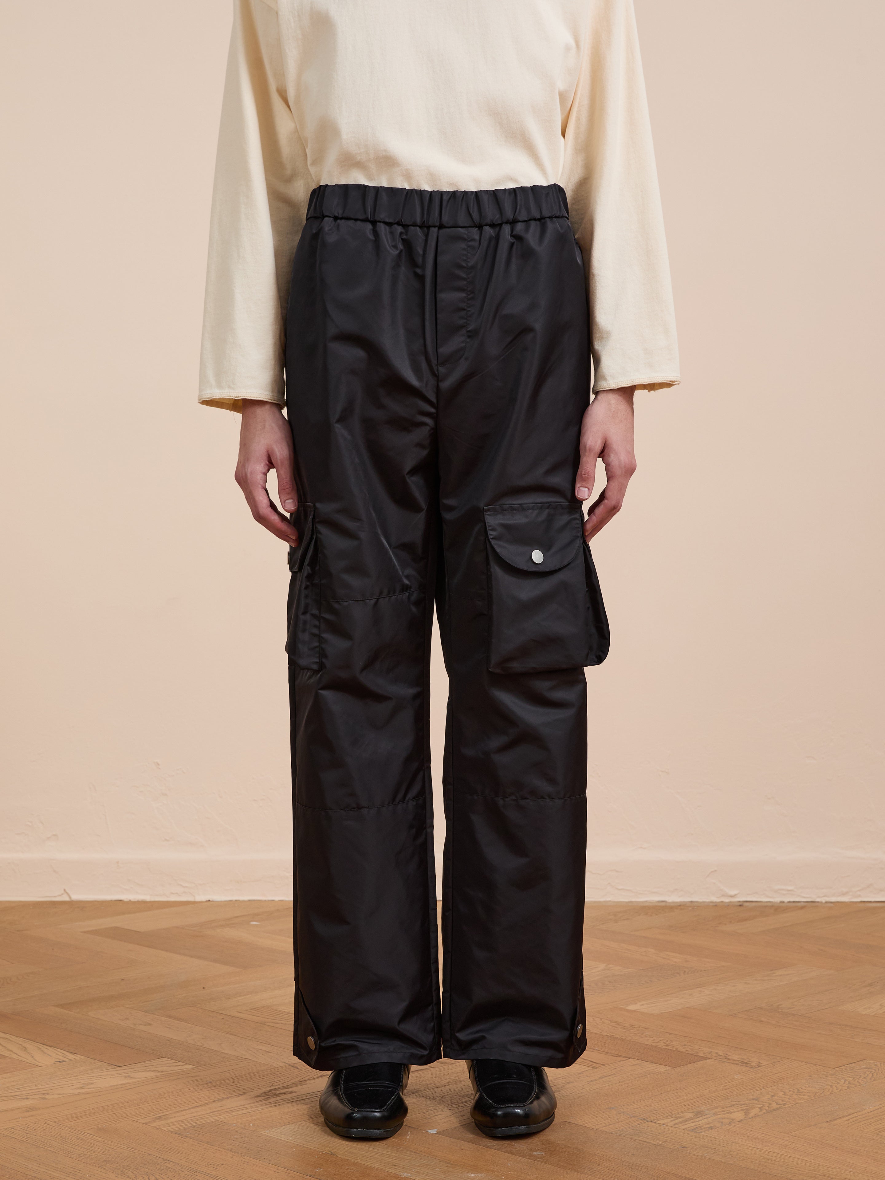 Wearing FOUND Nylon Cargo Pants with flap pockets and a relaxed fit, paired with a long-sleeve cream shirt and black shoes, a person stands on a wooden floor against a beige wall.