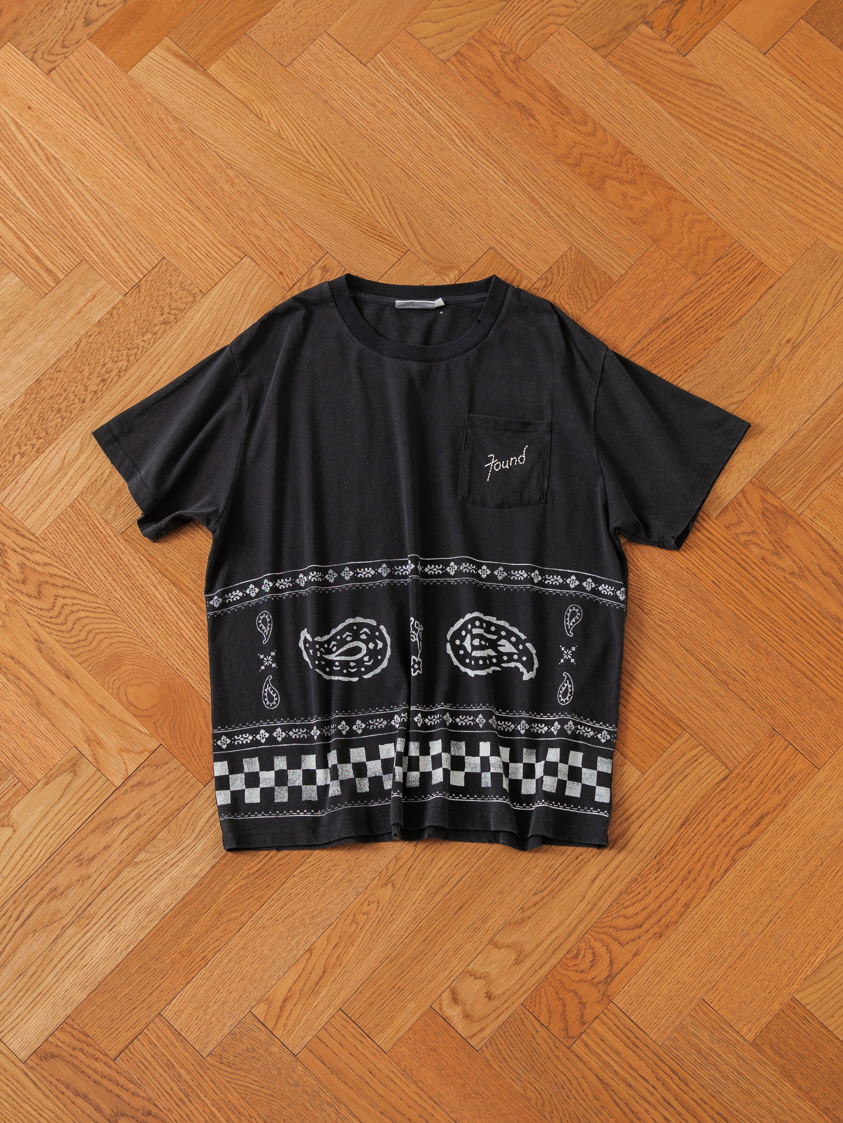 The Ornament Pocket Tee by FOUND, featuring white printed designs with Paisley patterns and checkered accents, is laid flat on a wooden parquet floor.