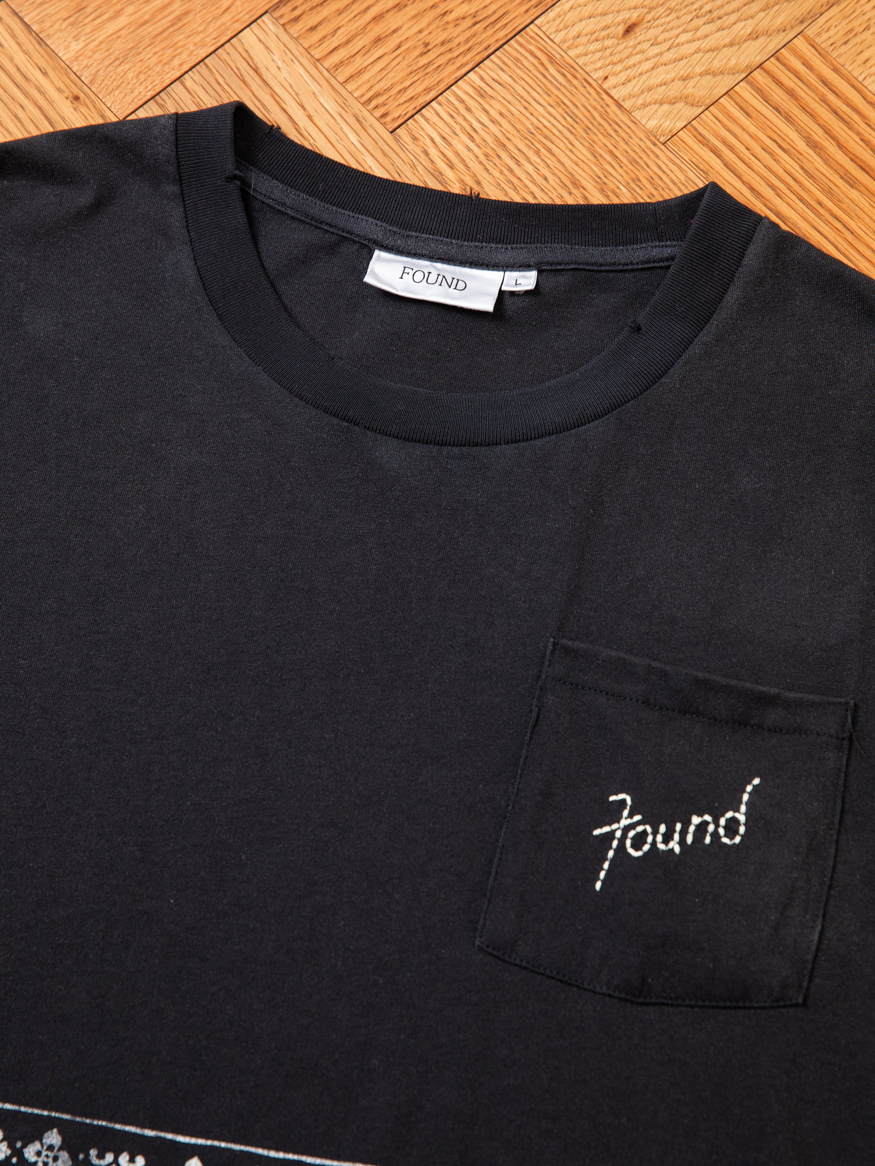 The Ornament Pocket Tee by FOUND is a black shirt featuring a small pocket on the left side of the chest. The word "FOUND" is embroidered on a tag inside the collar and also on the pocket. The shirt is displayed flat on a wooden surface.