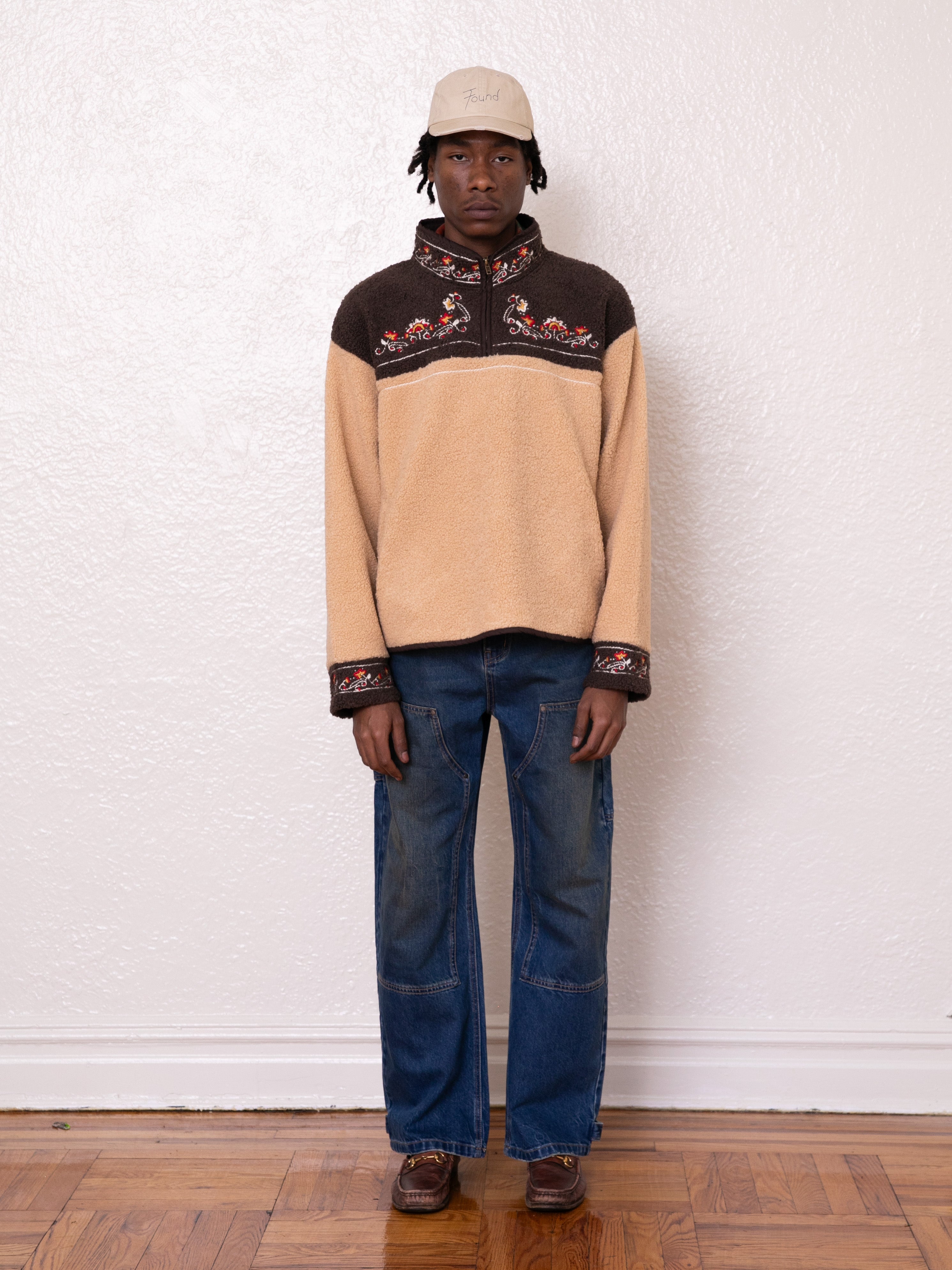 A person wearing the Pahari Floral Embroidered Sherpa Pullover by FOUND, paired with blue jeans, brown shoes, and a beige cap stands against a white wall on a wooden floor.