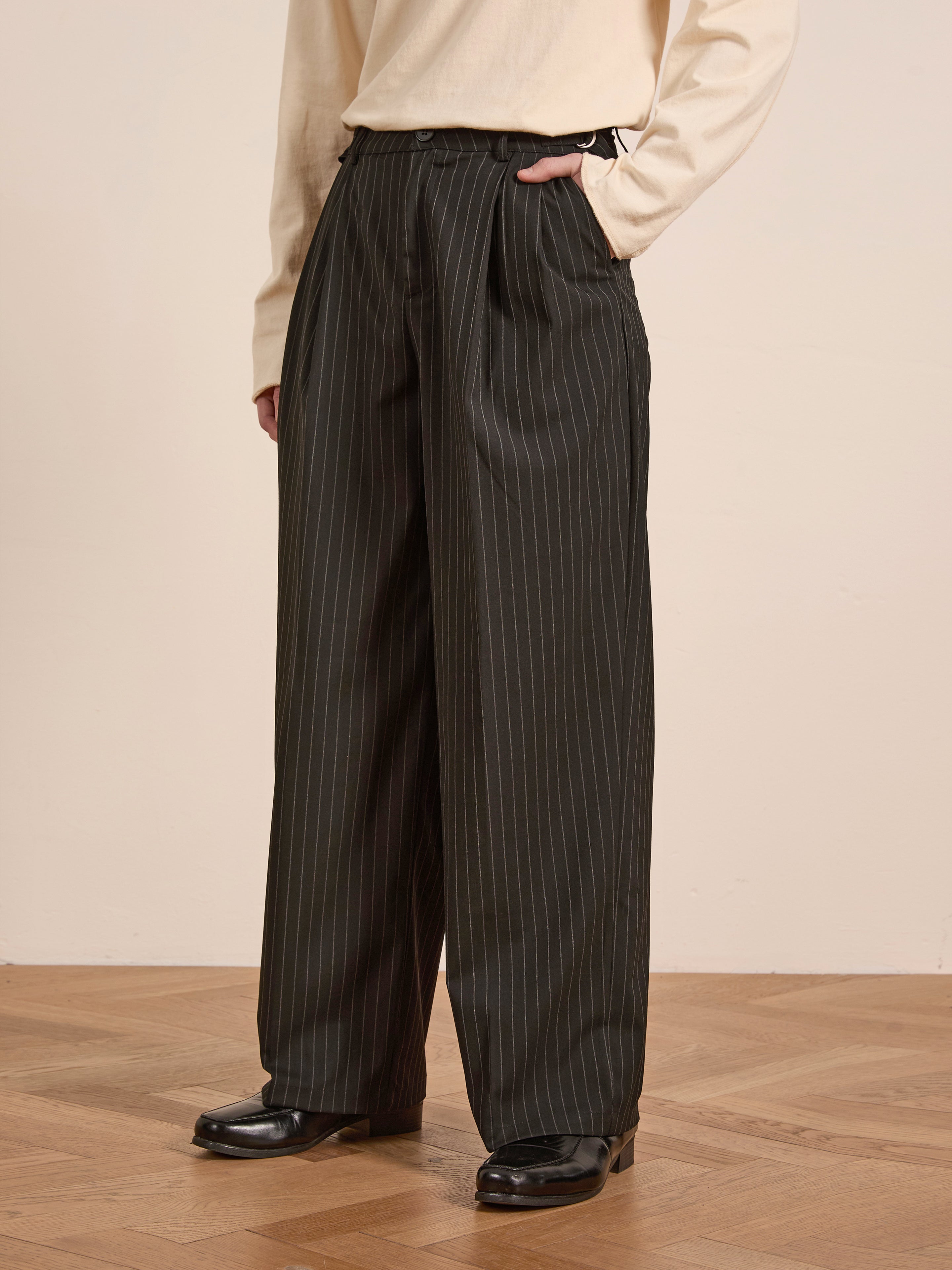 A person's lower half is visible, wearing FOUND's Pinstripe Pleated Trousers—dark pinstripe wide-leg pants with a relaxed fit—and a light-colored long-sleeve shirt, standing on a wooden floor.