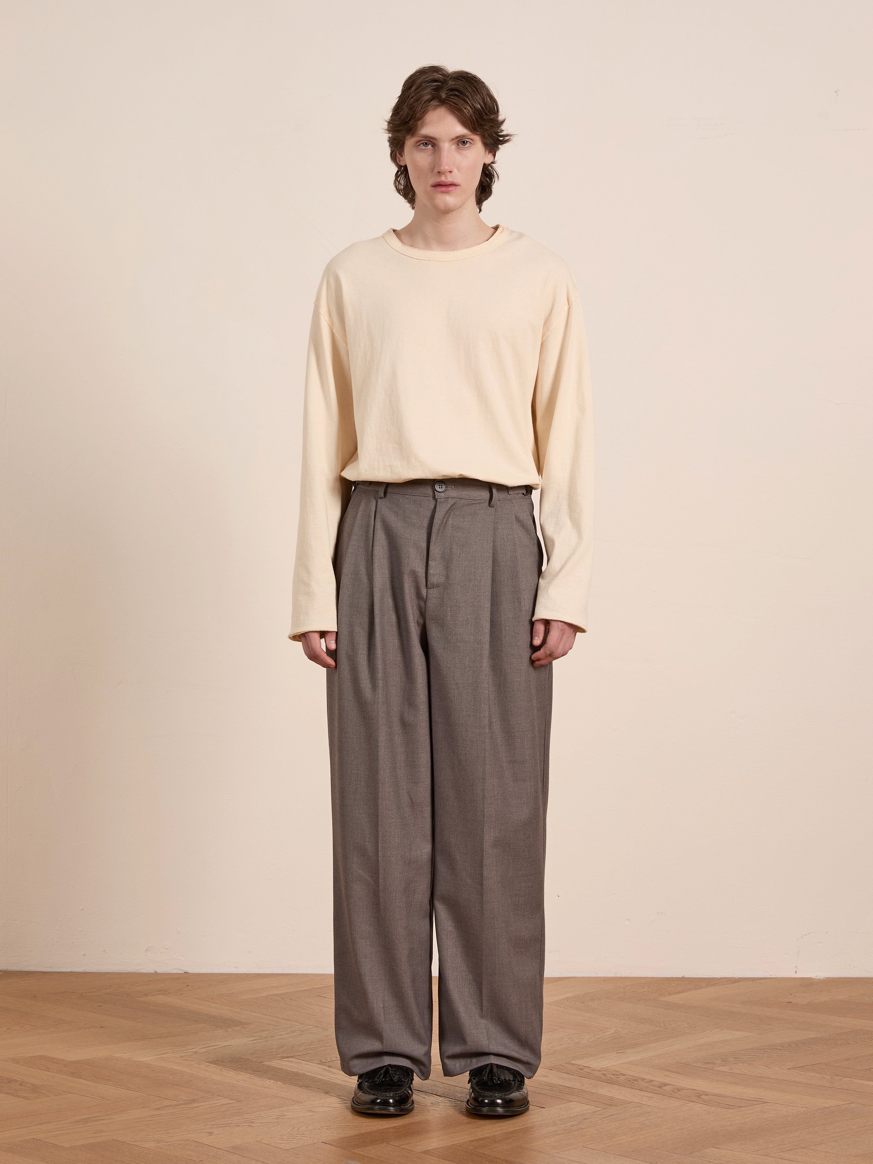 A person with short hair stands confidently in a cream long-sleeve shirt and sleek FOUND Pleated Trousers, featuring an adjustable waist and a high-rise double-pleat design. Black shoes complete the outfit against a plain beige backdrop.