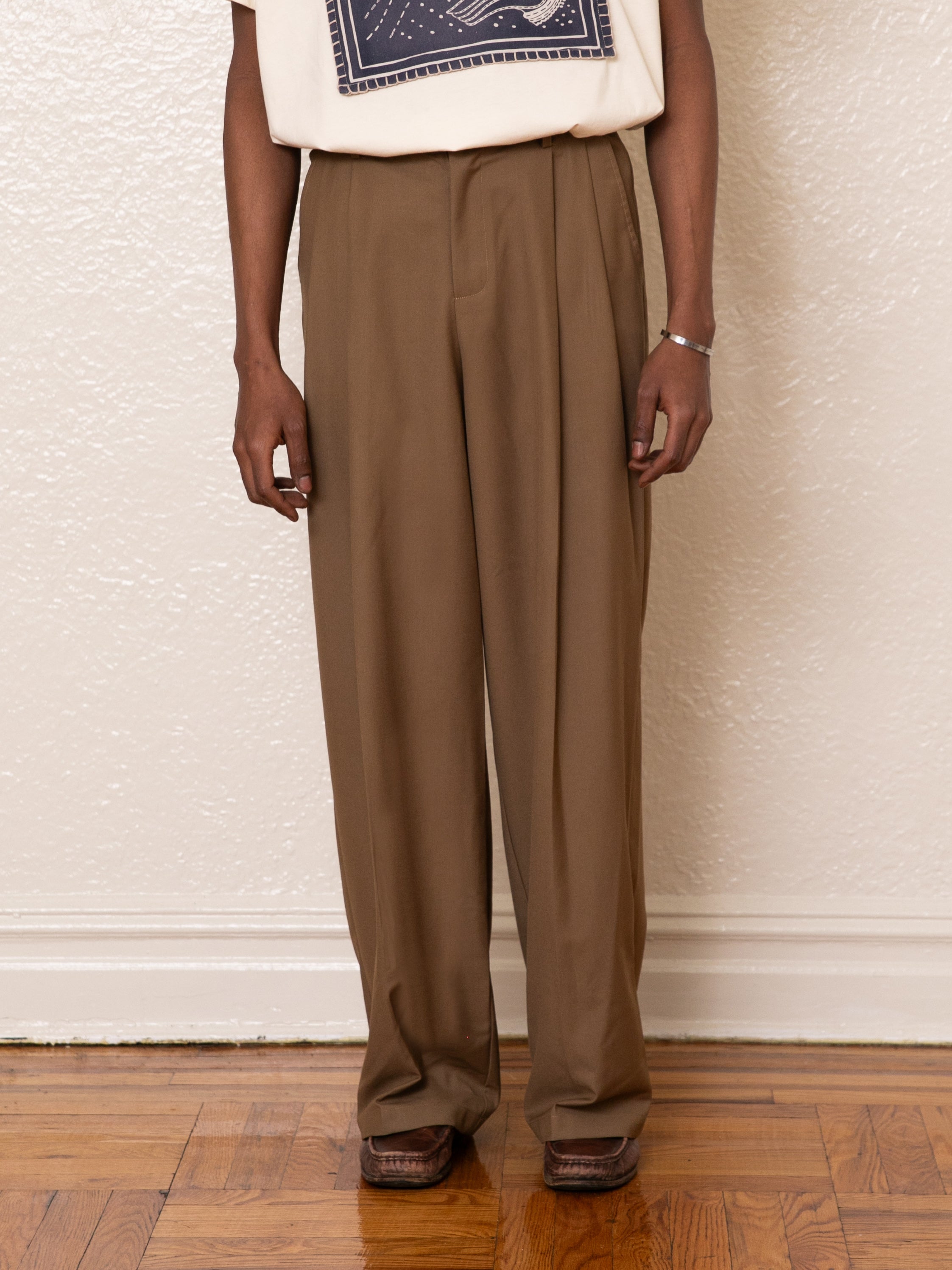 A person in FOUND Pleated Trousers stands against a white wall on a wooden floor, wearing a patterned top. Their relaxed arms emphasize the high-rise design's elegance.