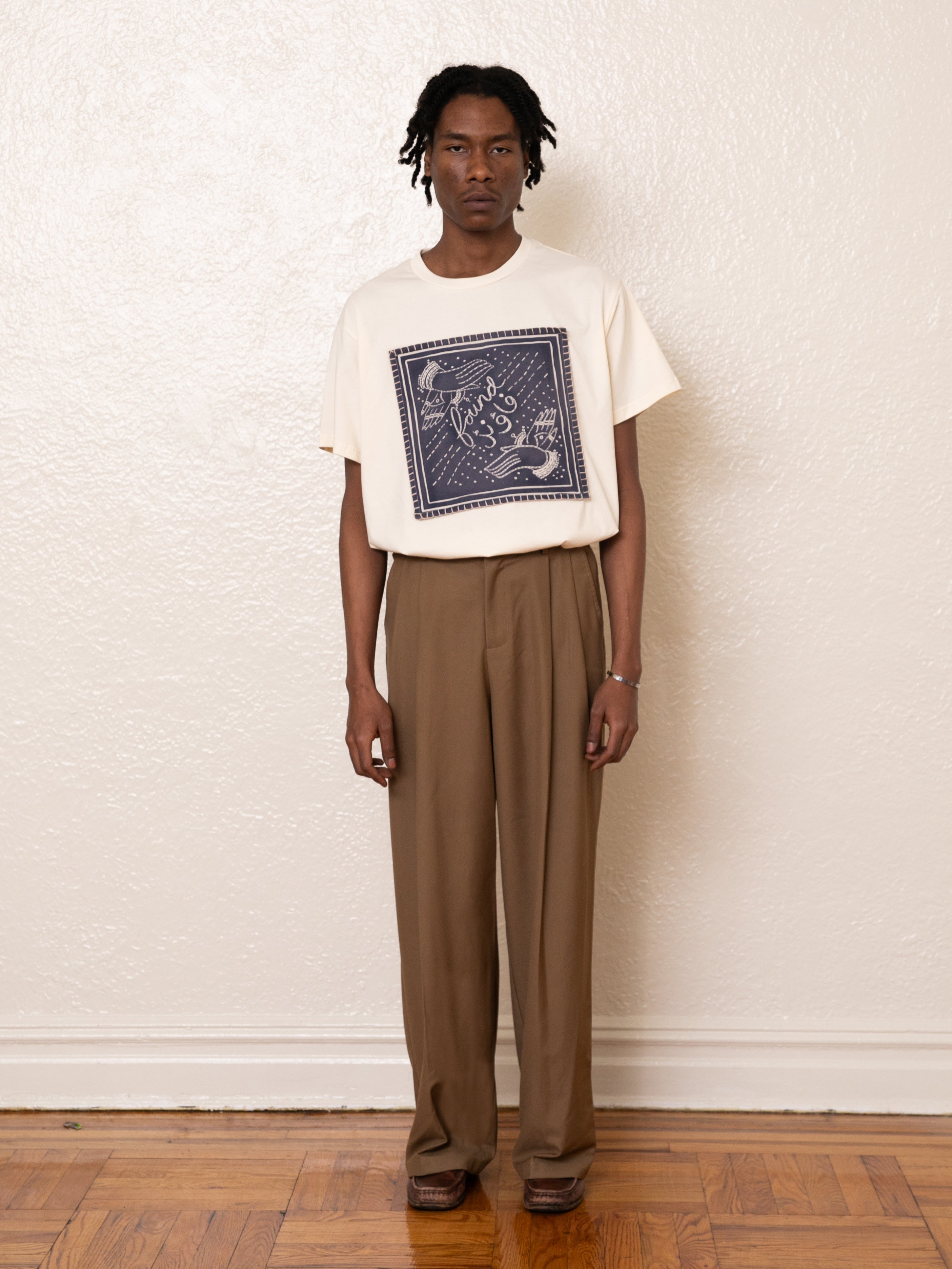 A person stands indoors wearing a white printed T-shirt and FOUND's Pleated Trousers in brown, featuring high-rise fit and adjustable waist tabs.